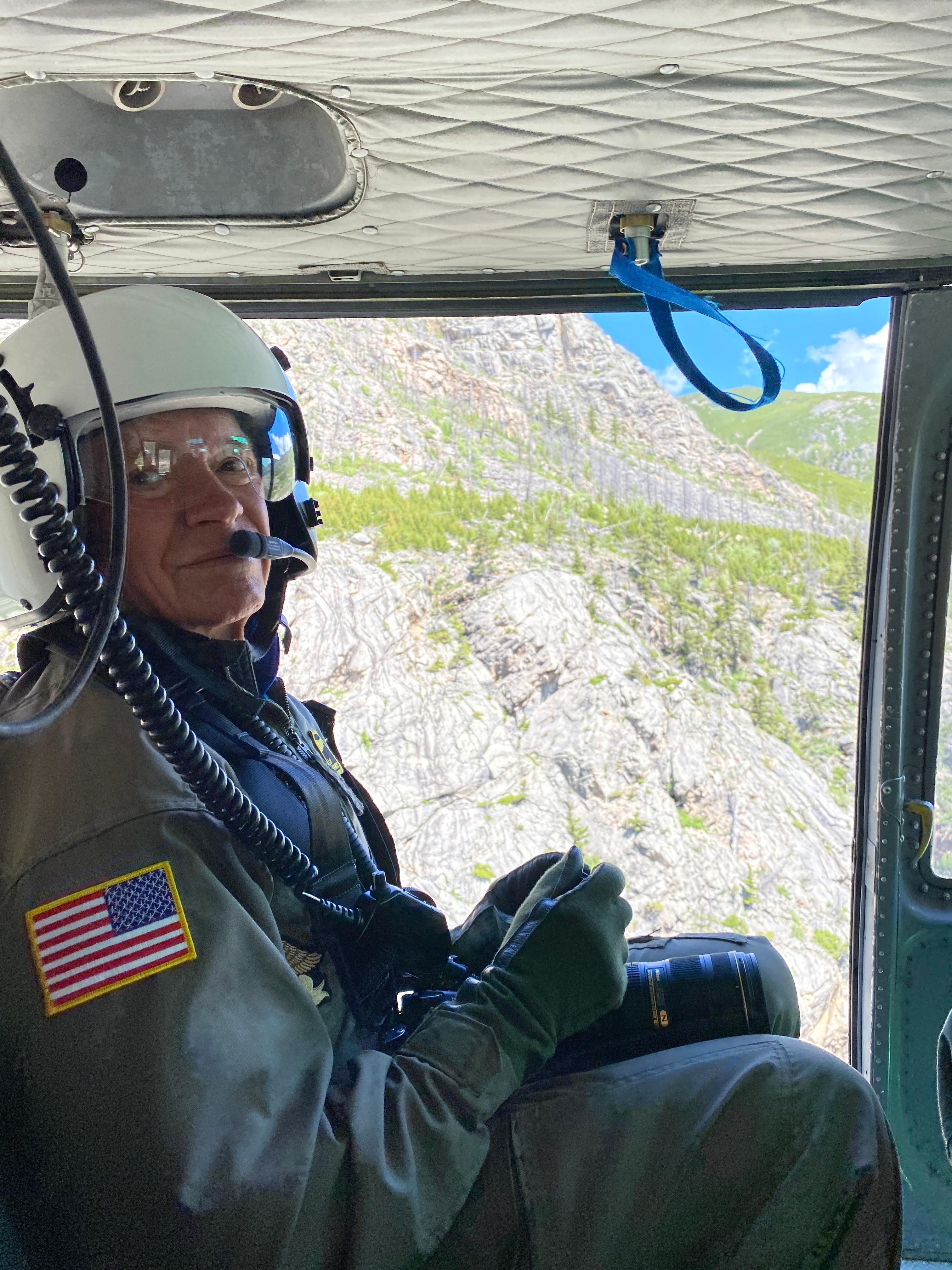 Man in green flight suit and white flight helmet seated screen left in a helicopter, mountains visible out of open door.