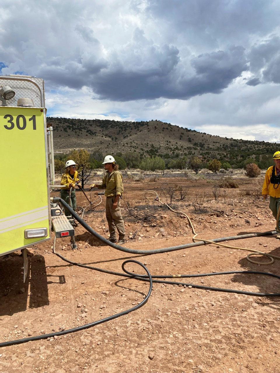 Fire crews in yellow shirts green pants white helmets, personal protective equipment talk logistics by a yellow wildland fire truck