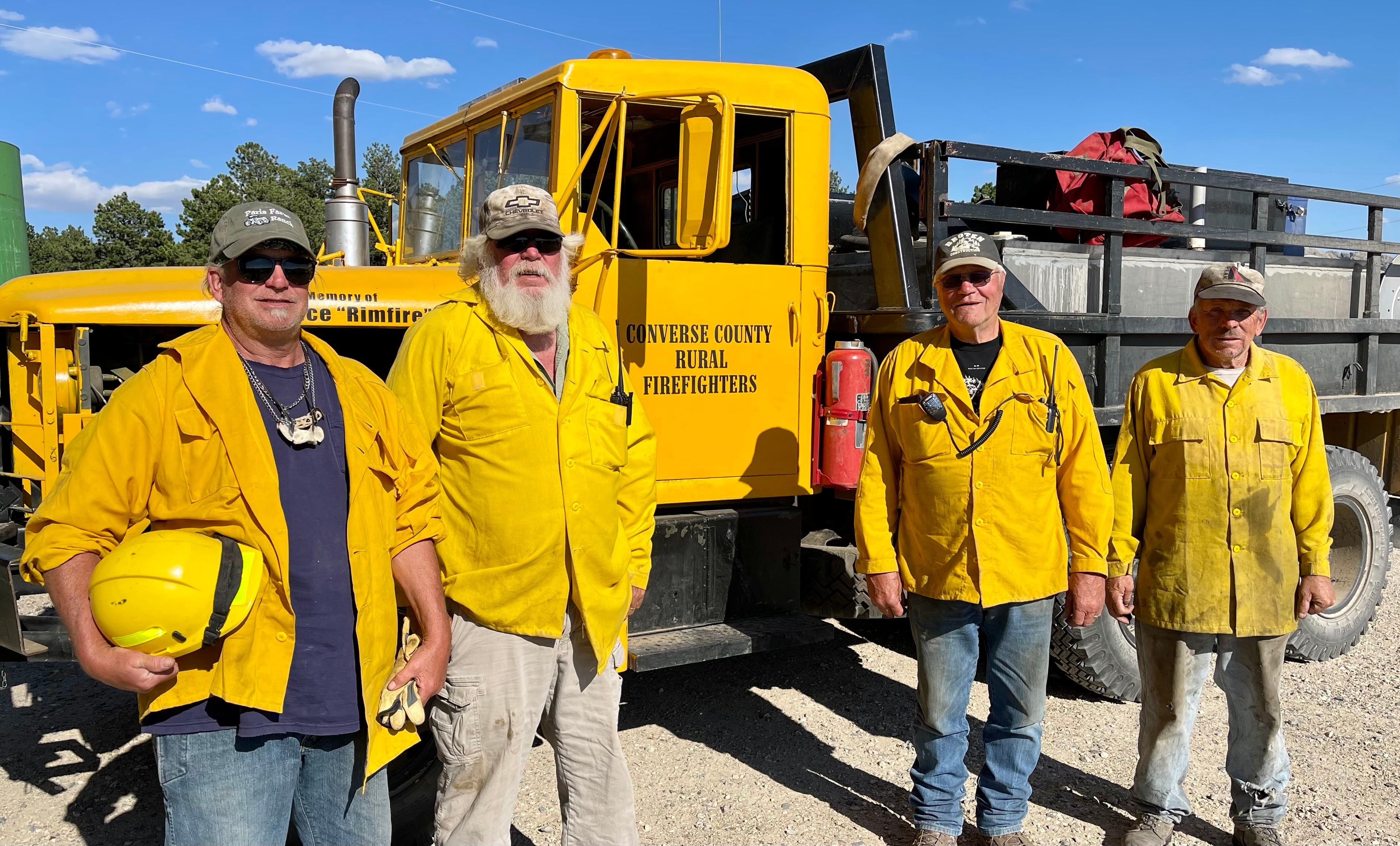 Four men wearing ball caps, yellow nomex shirts and blue jeans are standing in front of a yellow Converse County Rural Firefighters fire engine. 