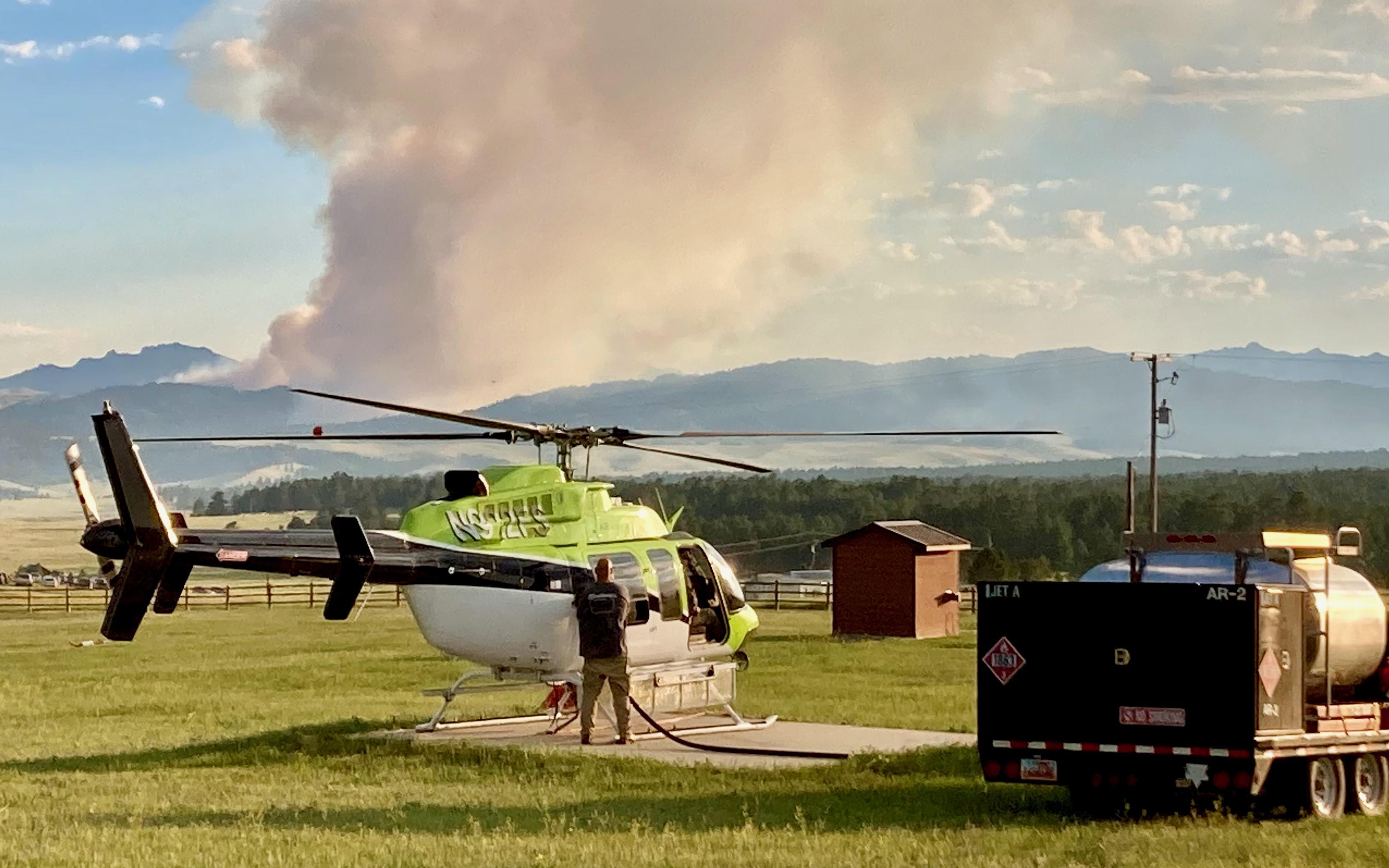 Green, black and white type one helicopter fuels up at Esterbrook Work Center, with a smoke column billowing up from the mountains in the background.