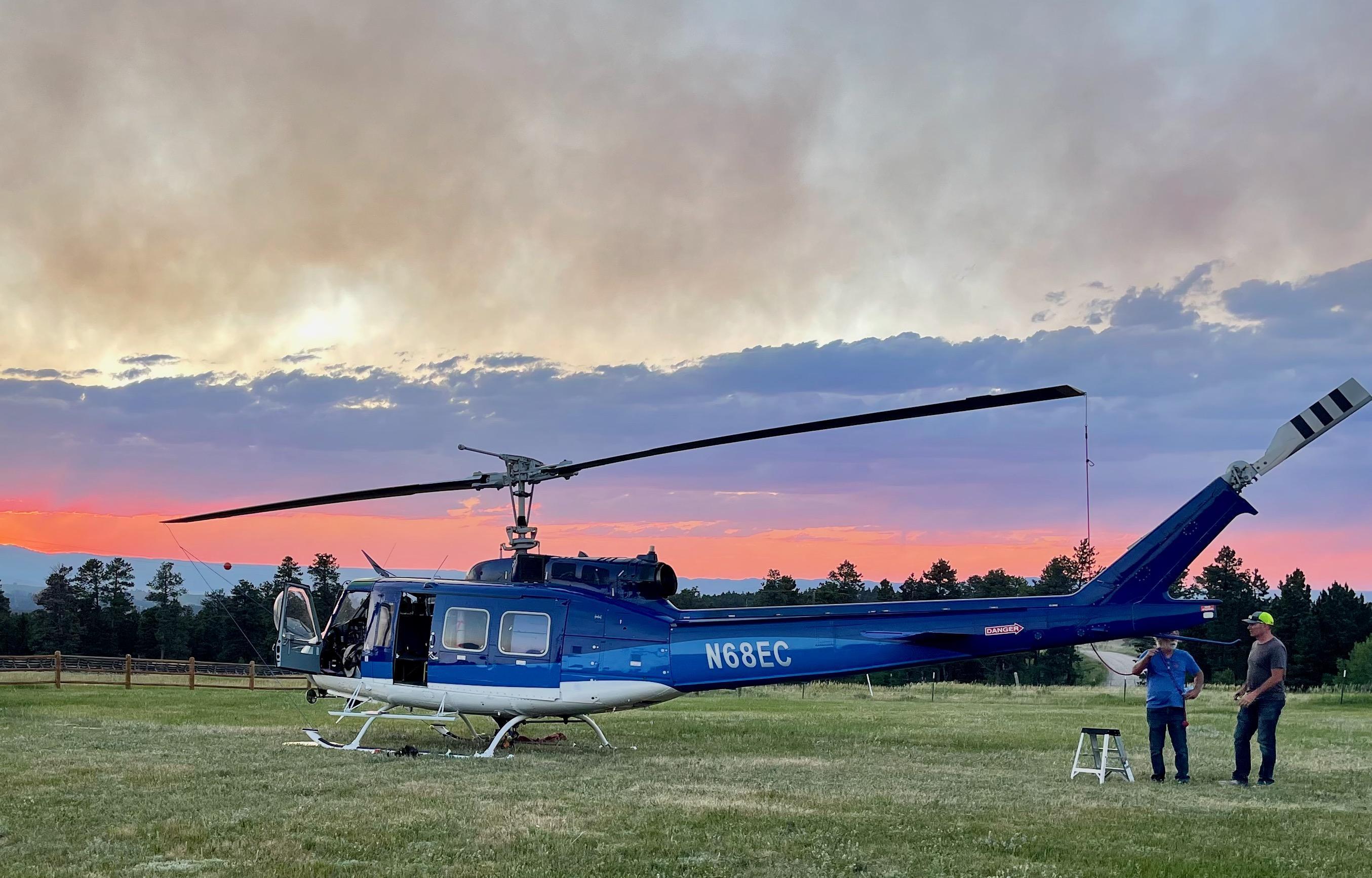 Blue and white Type 2 helicopter sits in the grass in front of a smoky pink and purple sunset