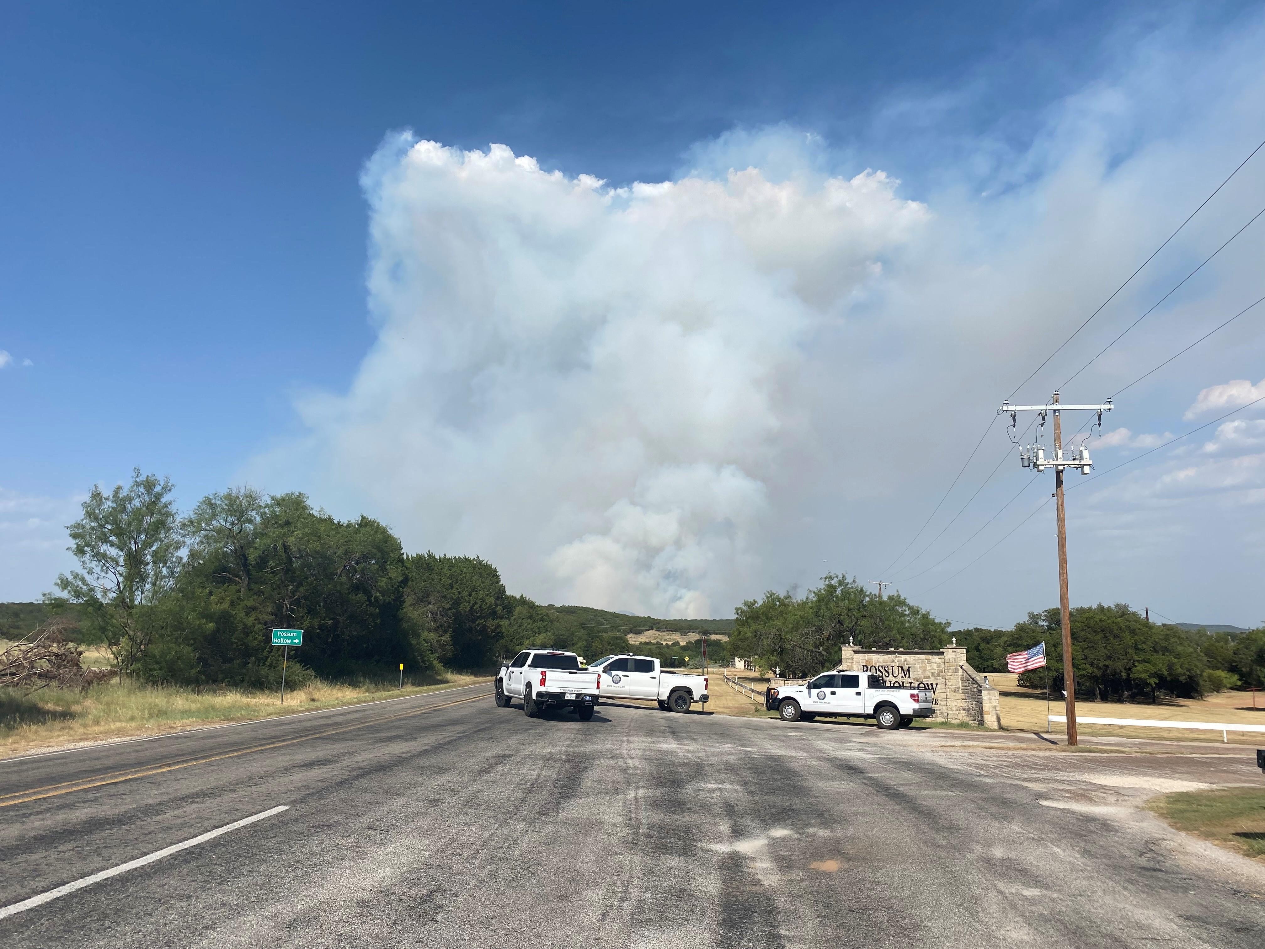 3 white trucks block the roadway leading to the 1148 Fire in Palo Pinto County. A smoke plume shows in the background with clear blue skies.