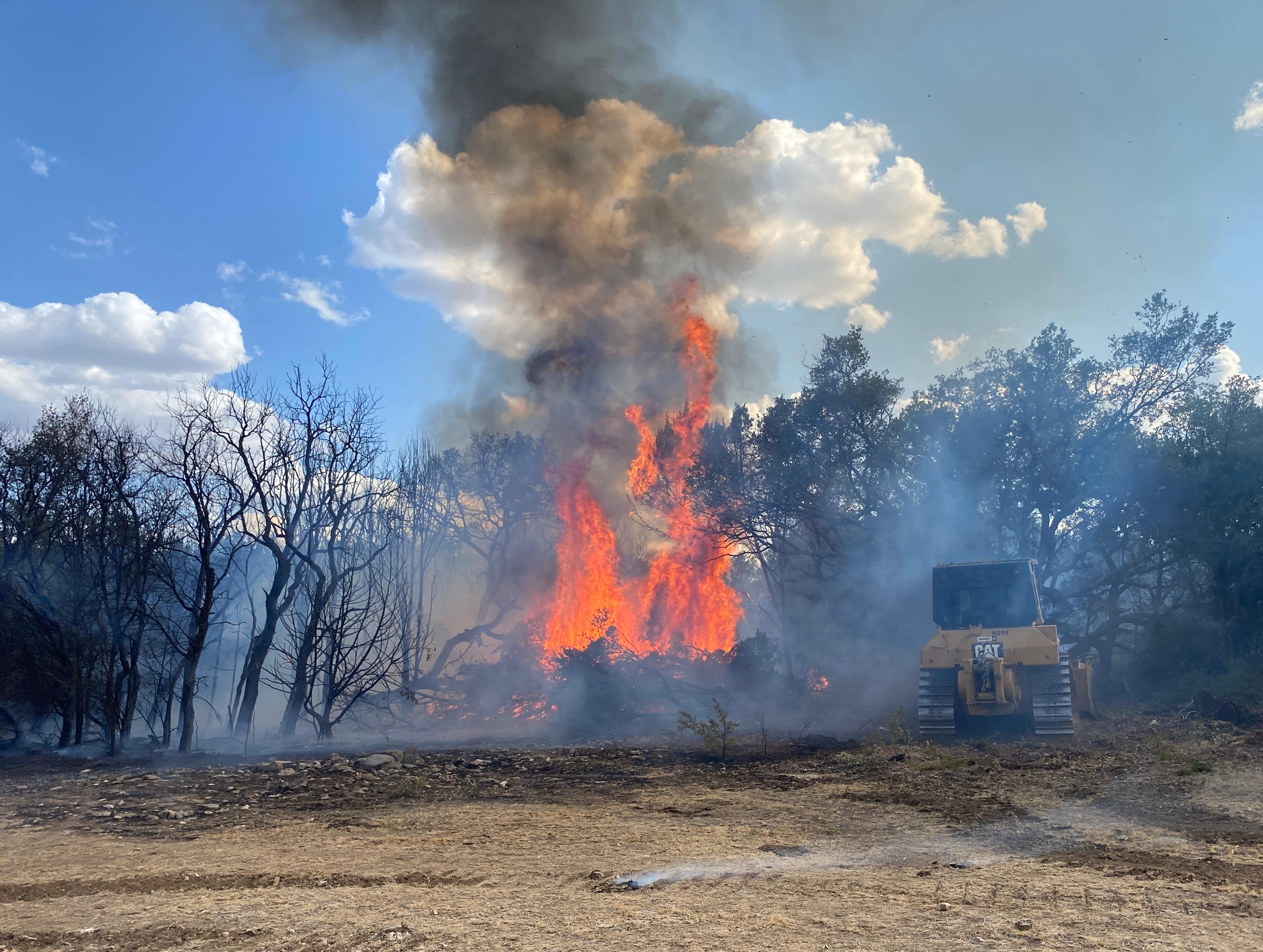 A yellow large bulldozer is next to a large plume of active red and orange flames. Many trees are in the area visibly smoking and charred. The sky is blue in the background with a plume of smoke