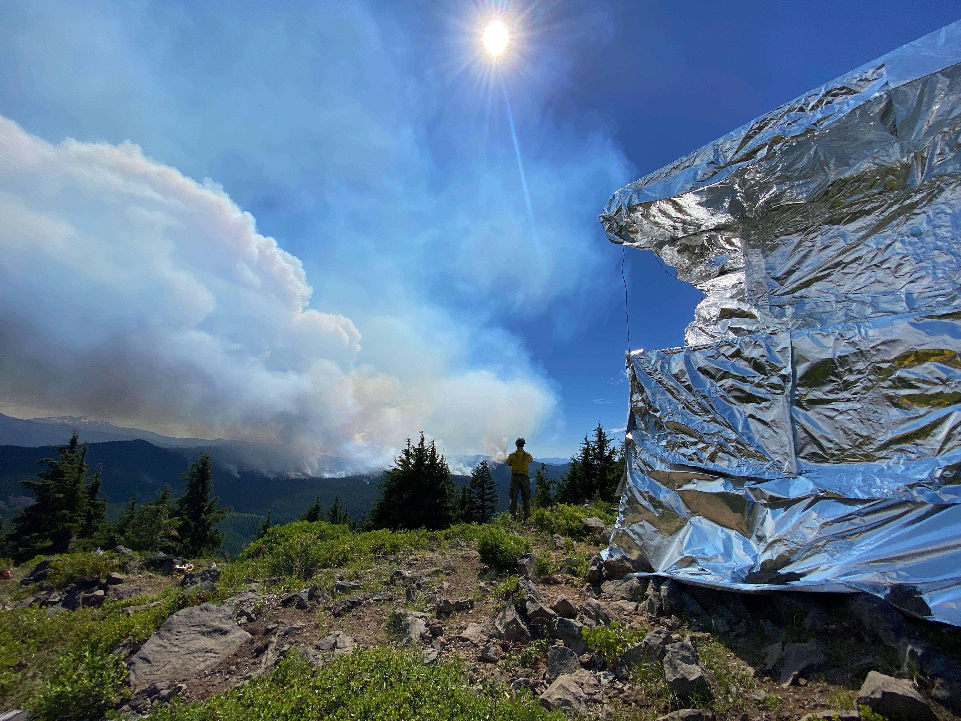 Waldo Mountain Lookout can be seen in the left corner of the frame with a reflective, aluminum wrap. A firefighter stands in center frame looking to the Southwest at the smoke being produced by the Cedar Creek Fire.