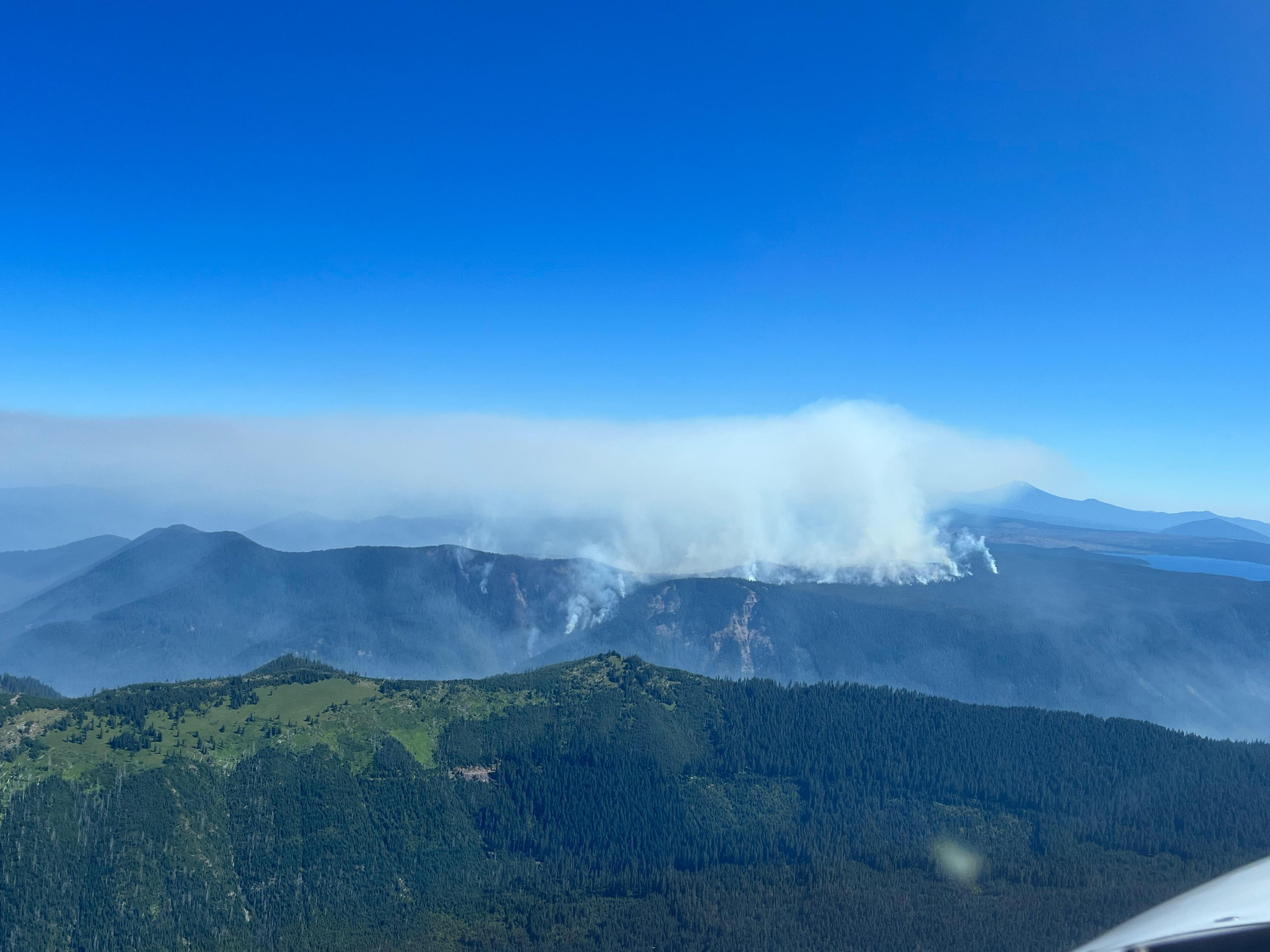 View of the smoke of Cedar Creek Fire from the south at approximately 11am on August 6. Smoke is dispersed across the fire area surrounded by dense timber. No column is visible.