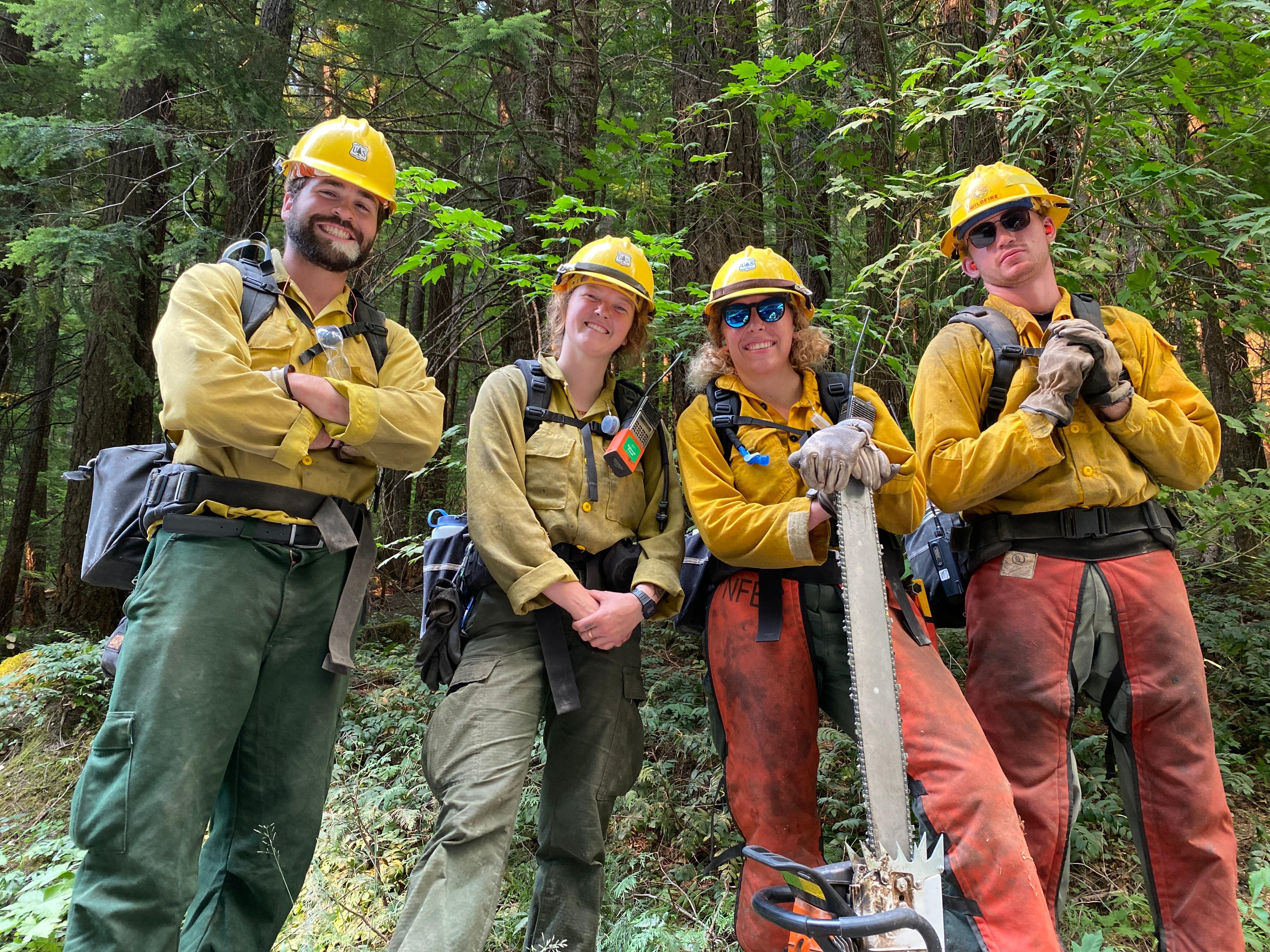 Two firefighters from an interagency crew from the Columbia River Gorge (BLM, USFS -GP -HOOD, and WA DNR) pose for a picture. One individual holds a large chainsaw, three others smiles holding their tools.