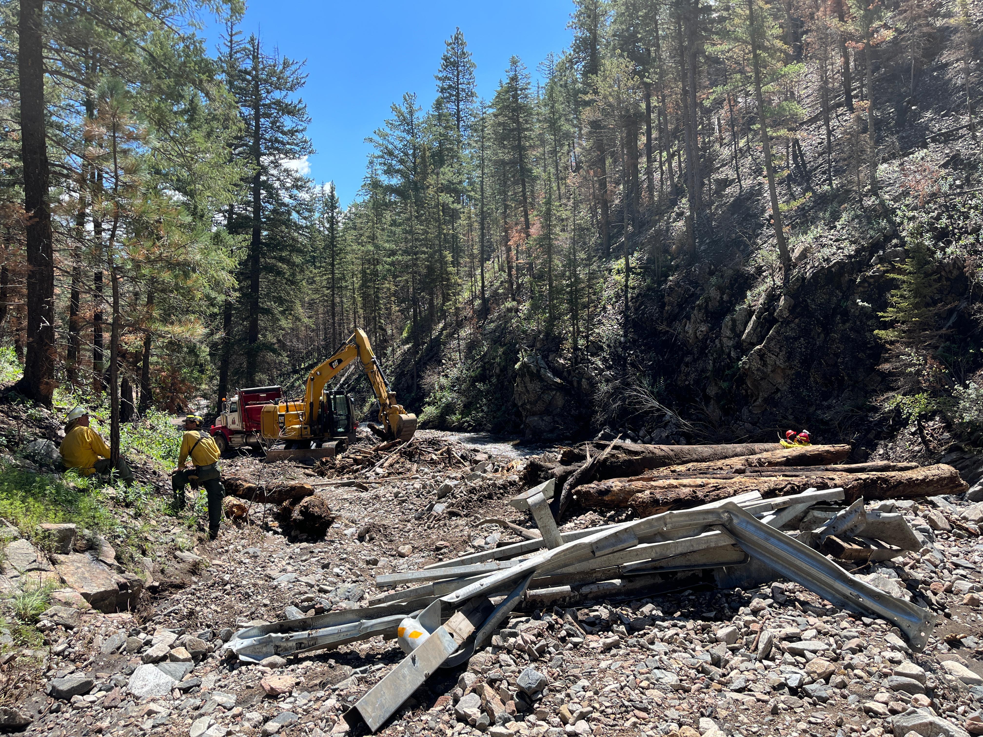 Image of flood debris piles of guardrails, rocks and logs being scooped up by an excavator on a road in the narrow bottom of canyon. Firefighters in safety gear are observing from a distance.