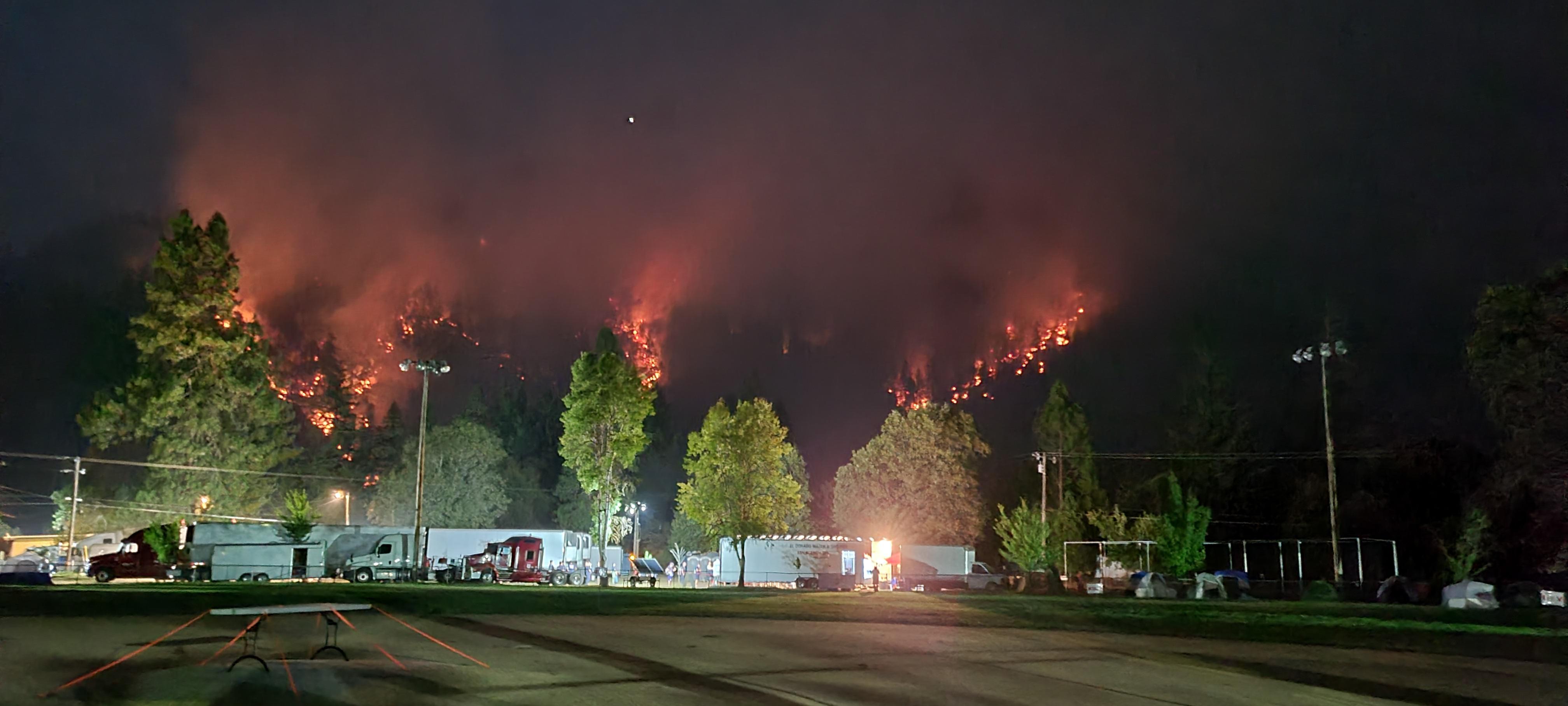 Photo depicts fire response support vehicles and trailers in a parking lot at the base of a steep forested slope. It's night and the dark trees on the slope are alight with fire glow.
