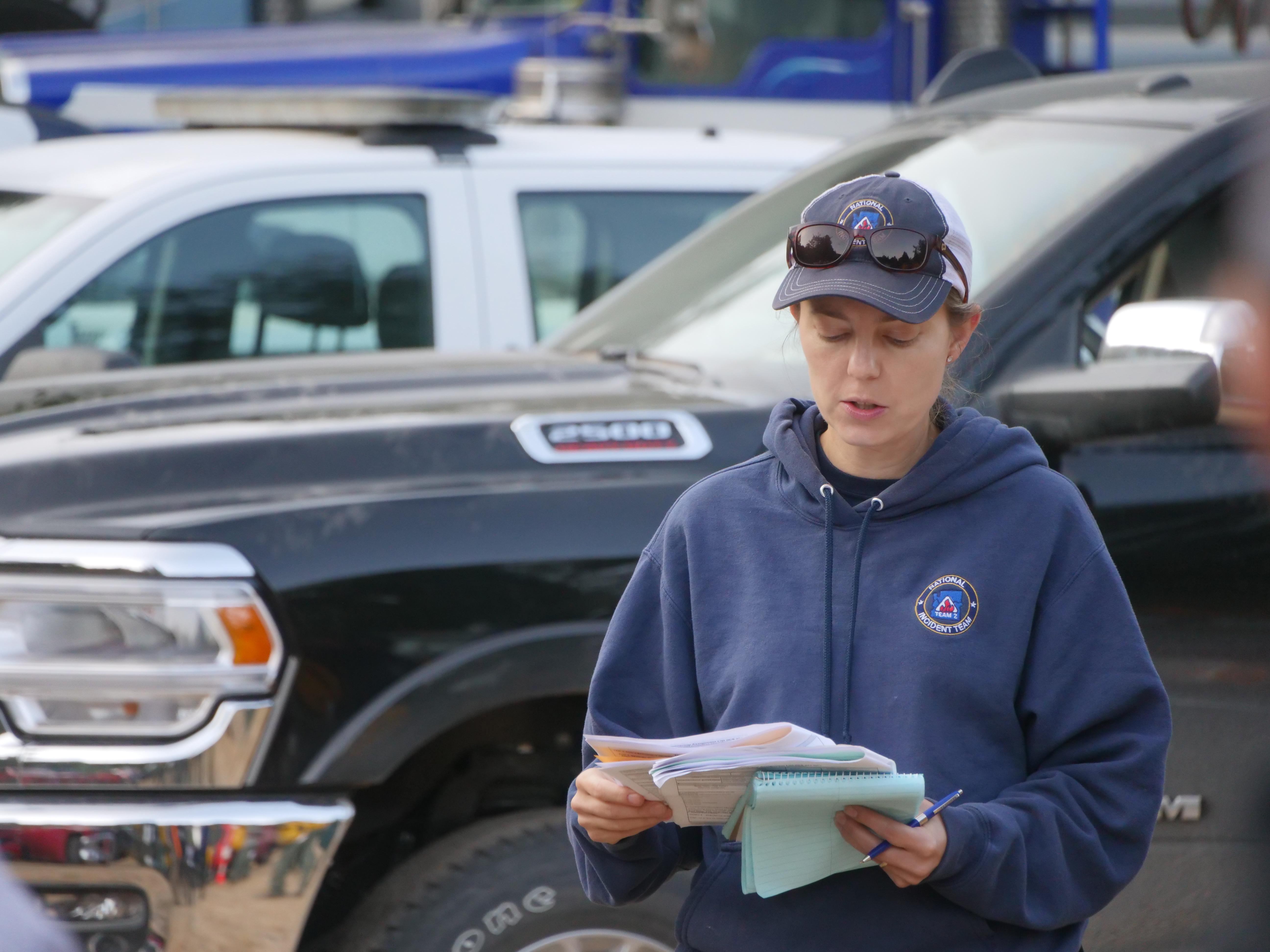 A fire supervisor is seen, looking at papers and a notebook, as she talks to other firefighters about their work assignments for the day.