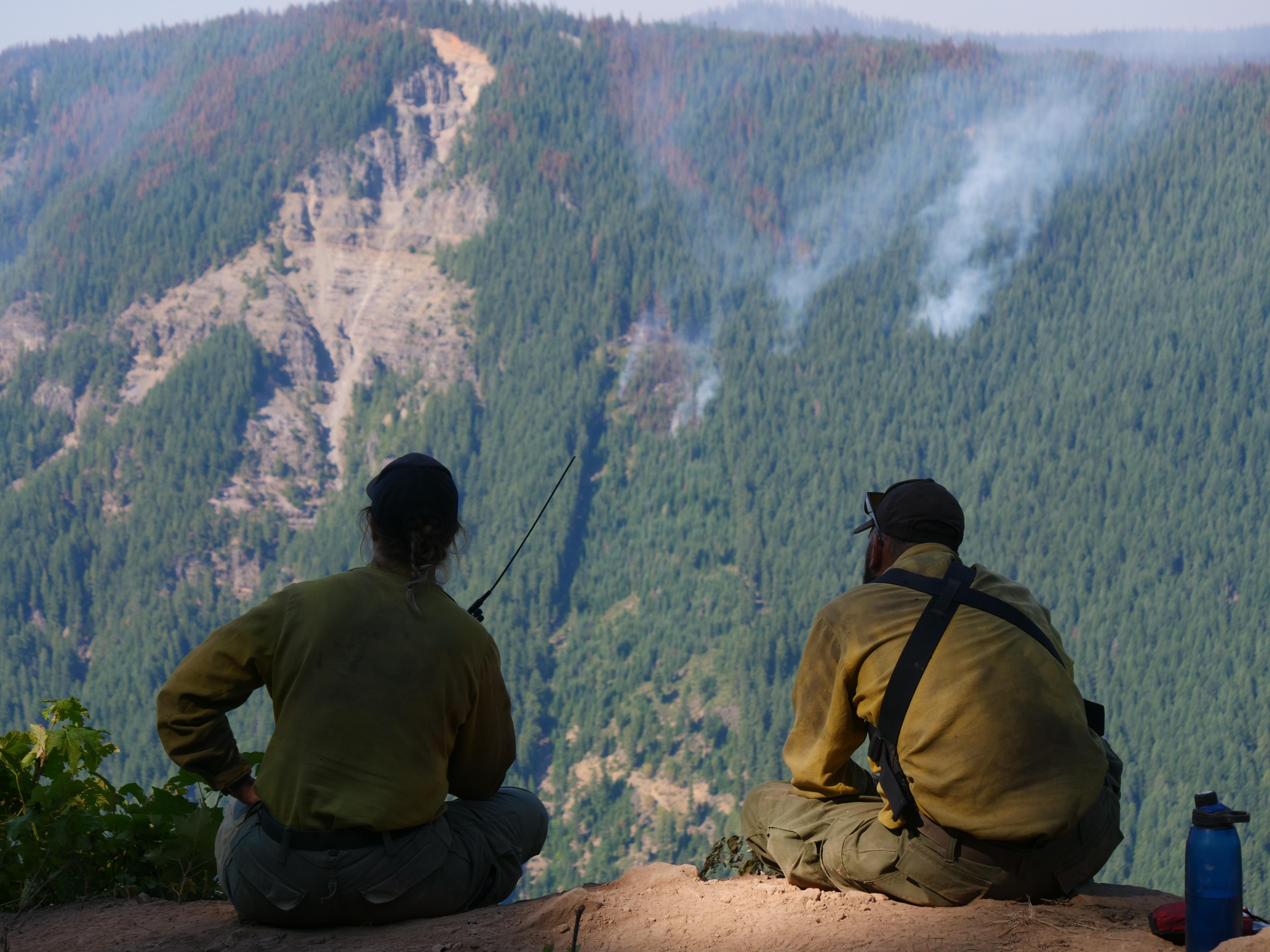 Two firefighters, one holding a radio, are seen sitting on a ridge across from a smoking hillside.