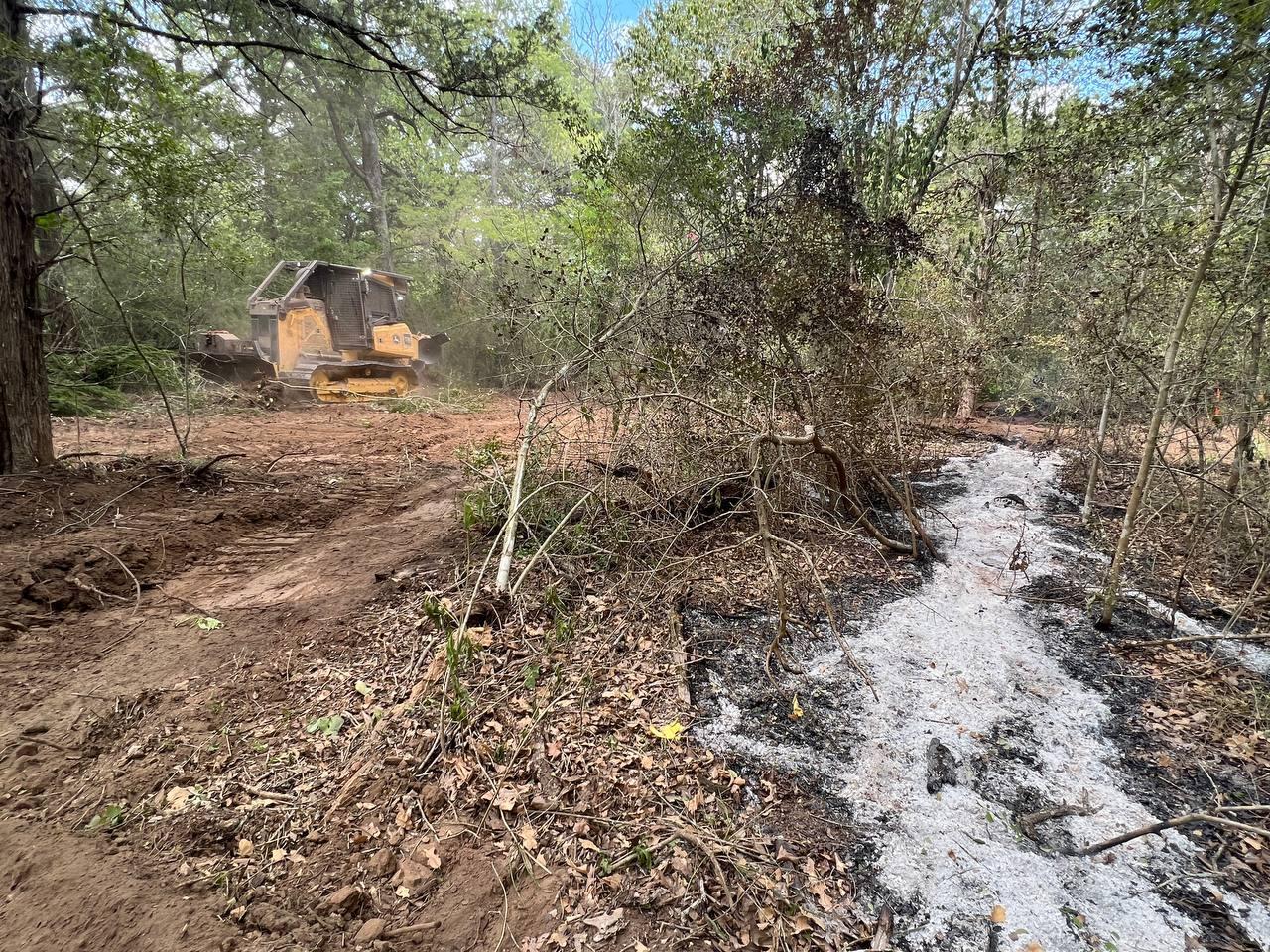 1 yellow piece of heavy equipment called a dozer works to scrape and push all vegetation away from a hotspot.  Hotspot is a long gray strip of gray ash that used to be a downed log.