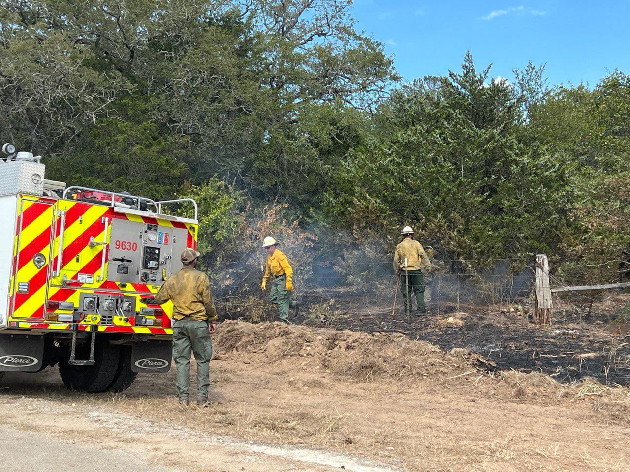 3 male firefighters with yellow shirts and green pants work around a fire engine with red and yellow stripes on the back.  Firefighter pulls fire hose through wire fence to put water on fire creeping underneath a tree.