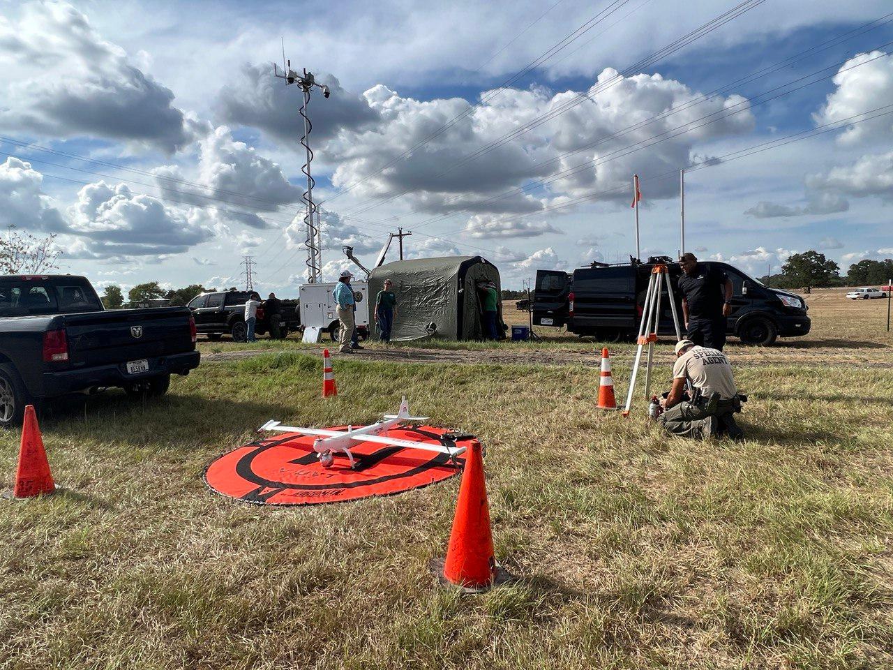 Multiple people working to set up equipment, tent, and drone launch pad to prepare for a recon flight.  Foreground has large, round orange mat on ground with a red and white drone set on top.