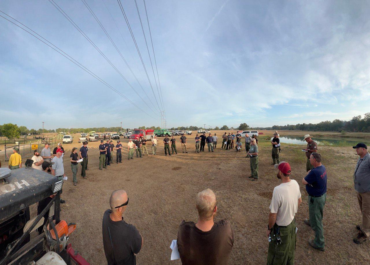 A couple dozen people stand in a circle in an open field to get information needed for the days operations.  Multiple fire vehicles are parked in the background with a cloudy blue sky.
