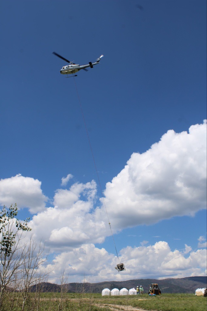 Image of a helicopter carrying  a spreading bucket full of seed away from Johnson Mesa. Crew and large white bags of seed are under the helicopter. 