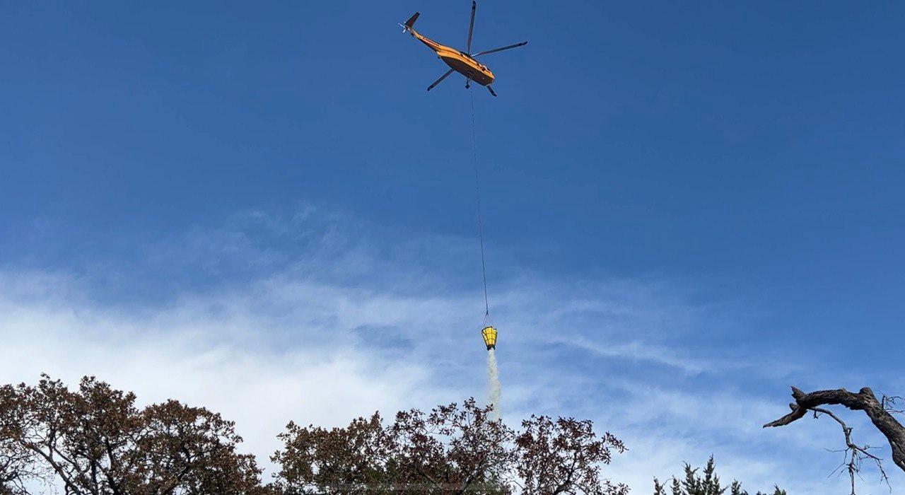 Yellow helicopter with a long line holding a water bucket. Water is being dumped out of bucket and onto trees below. Sky is partly cloud with blue.