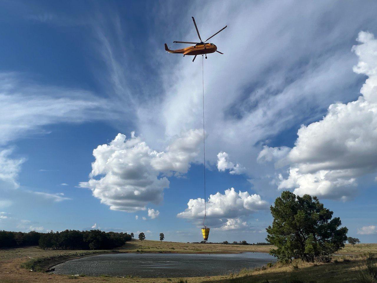 A yellow helicopter hovers over a pond. A long line hangs from the helo and has a yellow bucket at the end. Partly cloudy blue sky in background.