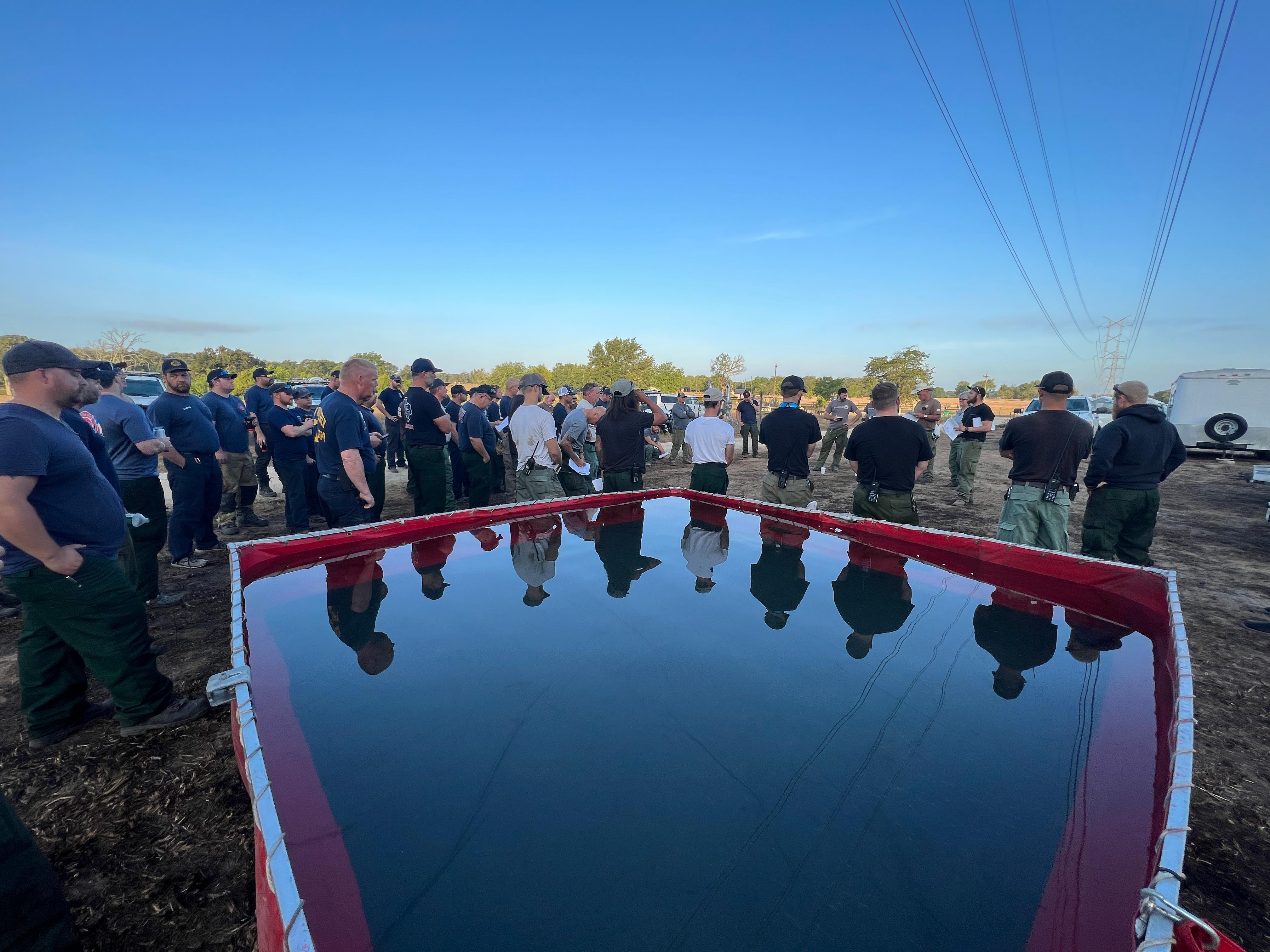 Approximately 60 people stand in a semi-circle.  Foreground has square, red tank filled with water casting a reflection of those standing nearby.