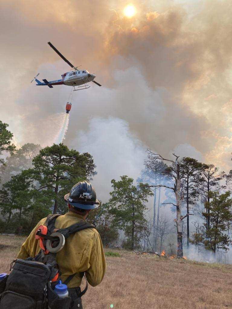 Smokey filled sky with fire along ground in trees and grass. Firefighter stands in foreground while a helicopter drops water from a hanging bucket onto the fire.