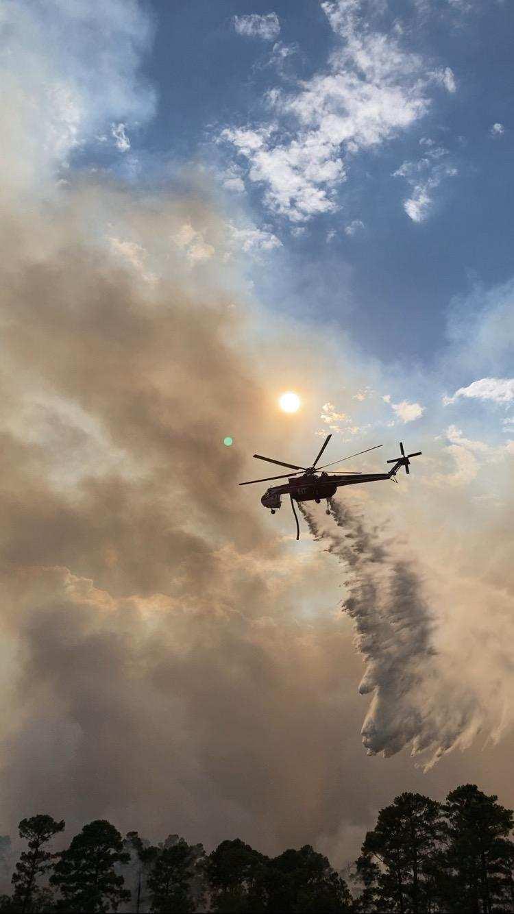 Smoke rises above tree line against cloudy, blue sky. Helicopter drops water that falls towards the ground.