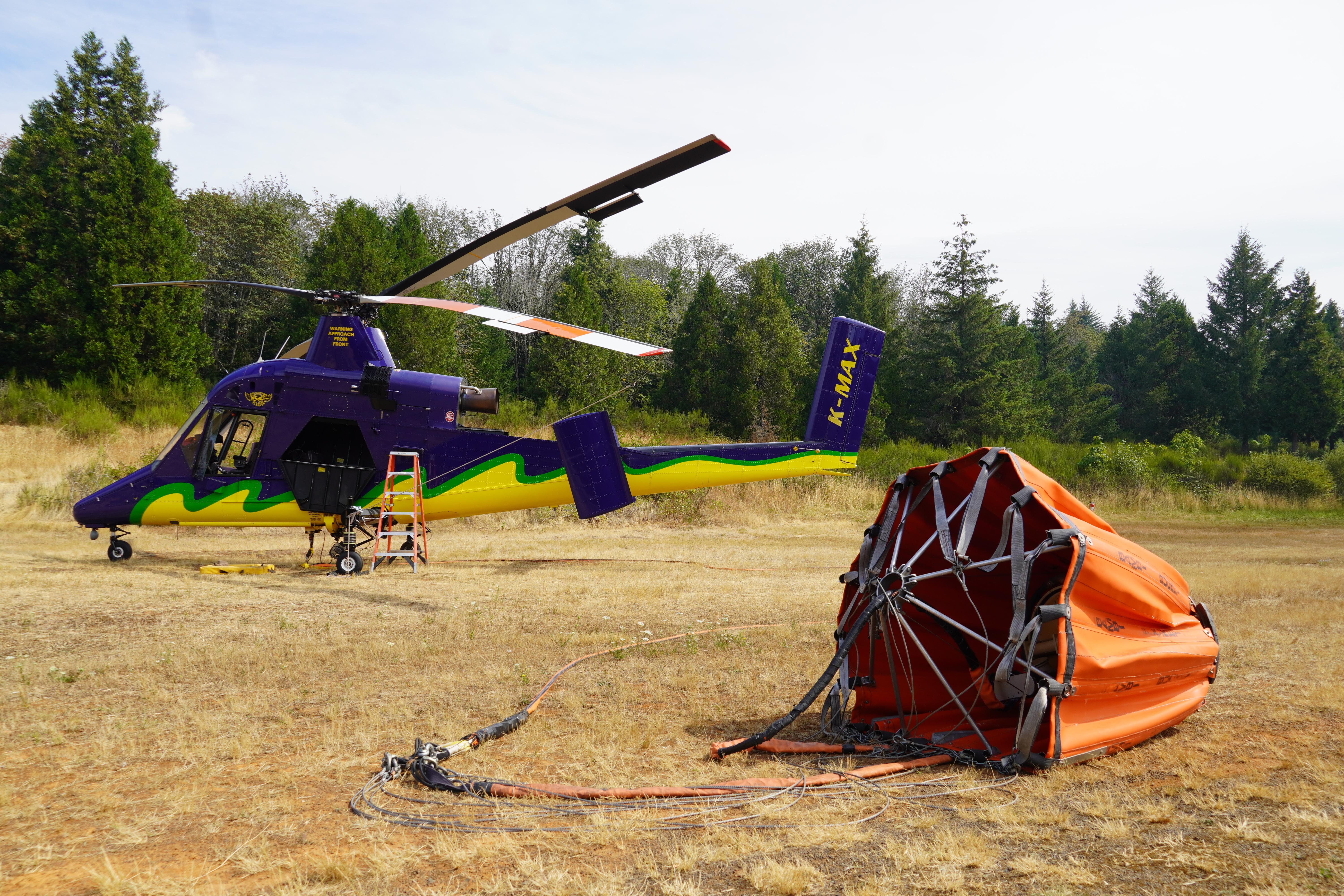 A large helicopter, painted purple and yellow with a green border between those two colors is seen sitting stationary on the ground. Its water bucket, connected by a large cable, is shown in the foreground.