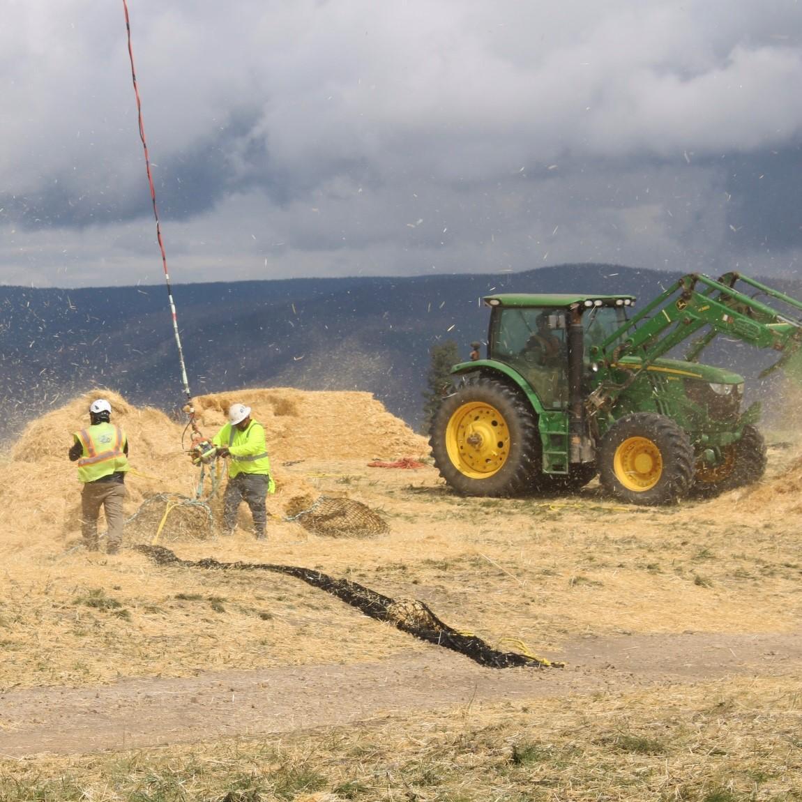 Two men work to fill a large net  hanging from a helicopter full of straw. There is a tractor to the right of them moving bales of straw.