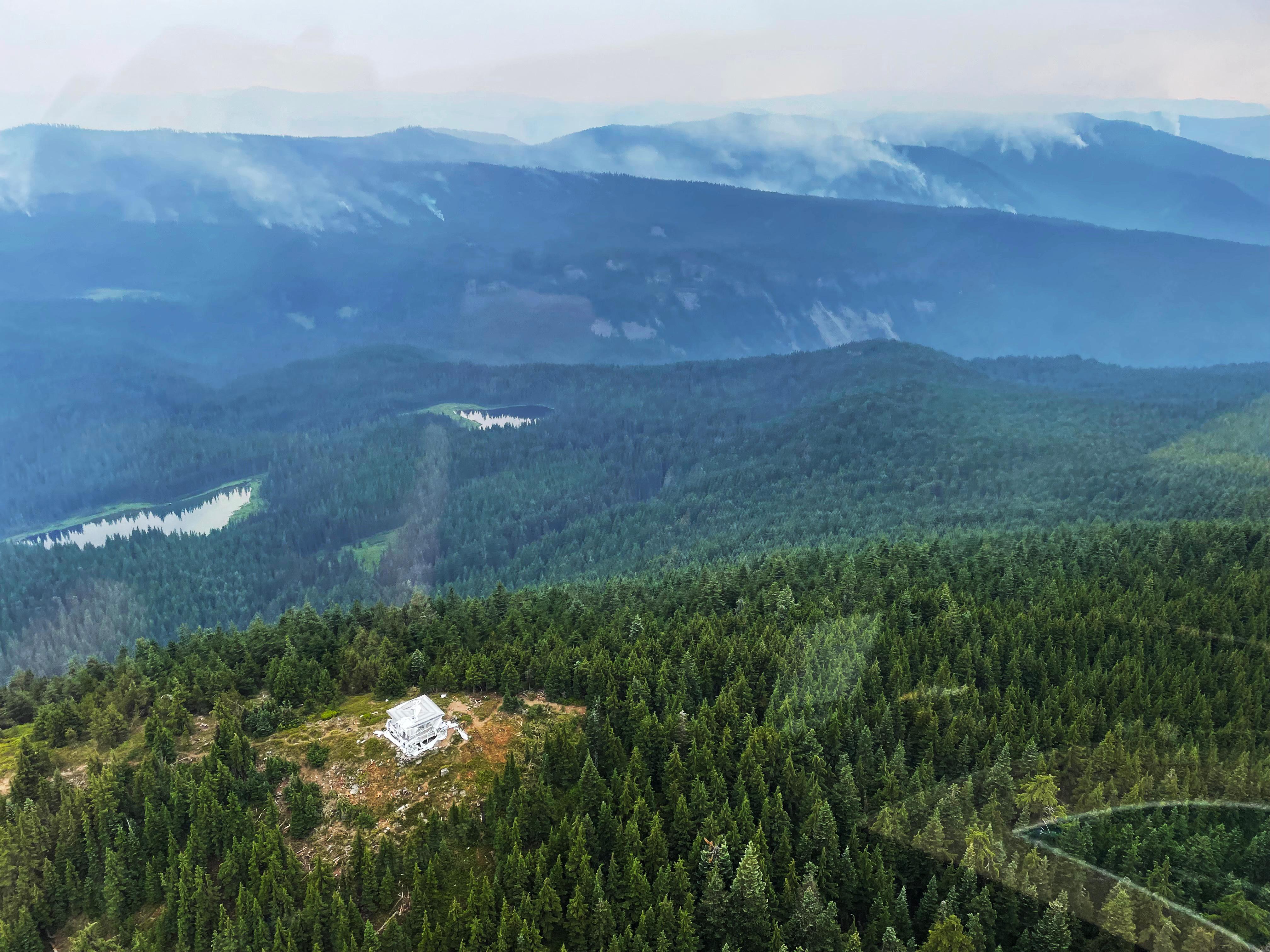 An aerial photograph showing fire lookout tower wrapped in protective foil. Smoke can be seen drifting up from many places in the forested mountains in the distance.