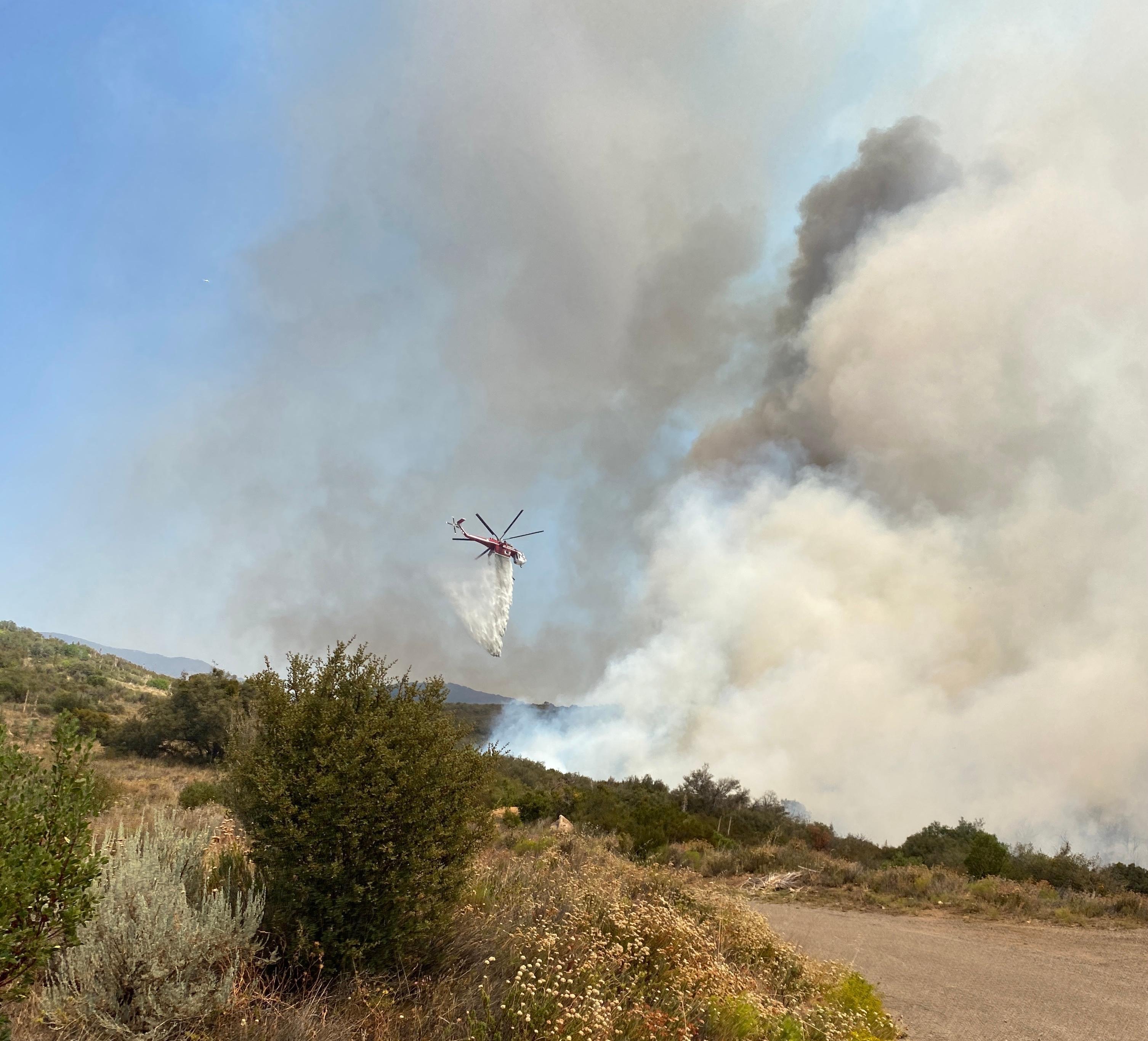Large helicopter (Type one) dropping water on a section of the fire, smoke and brush in the foreground