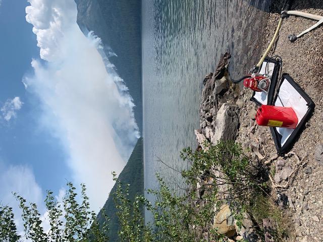 Smoke rises from the Quartz fire in the distance with Quartz lake midground and a pump and fuel can on the shore in the foreground.