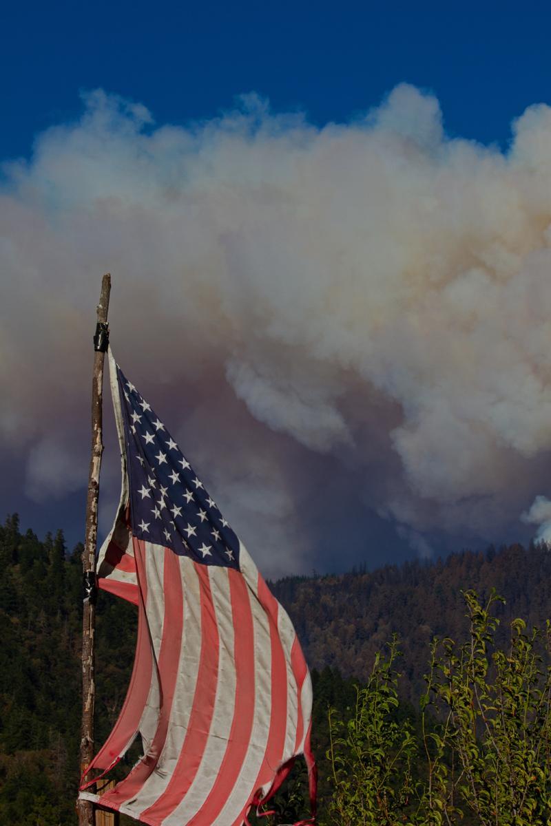 An American Flag attached to a stick in front of a smoke plume rising above mountains