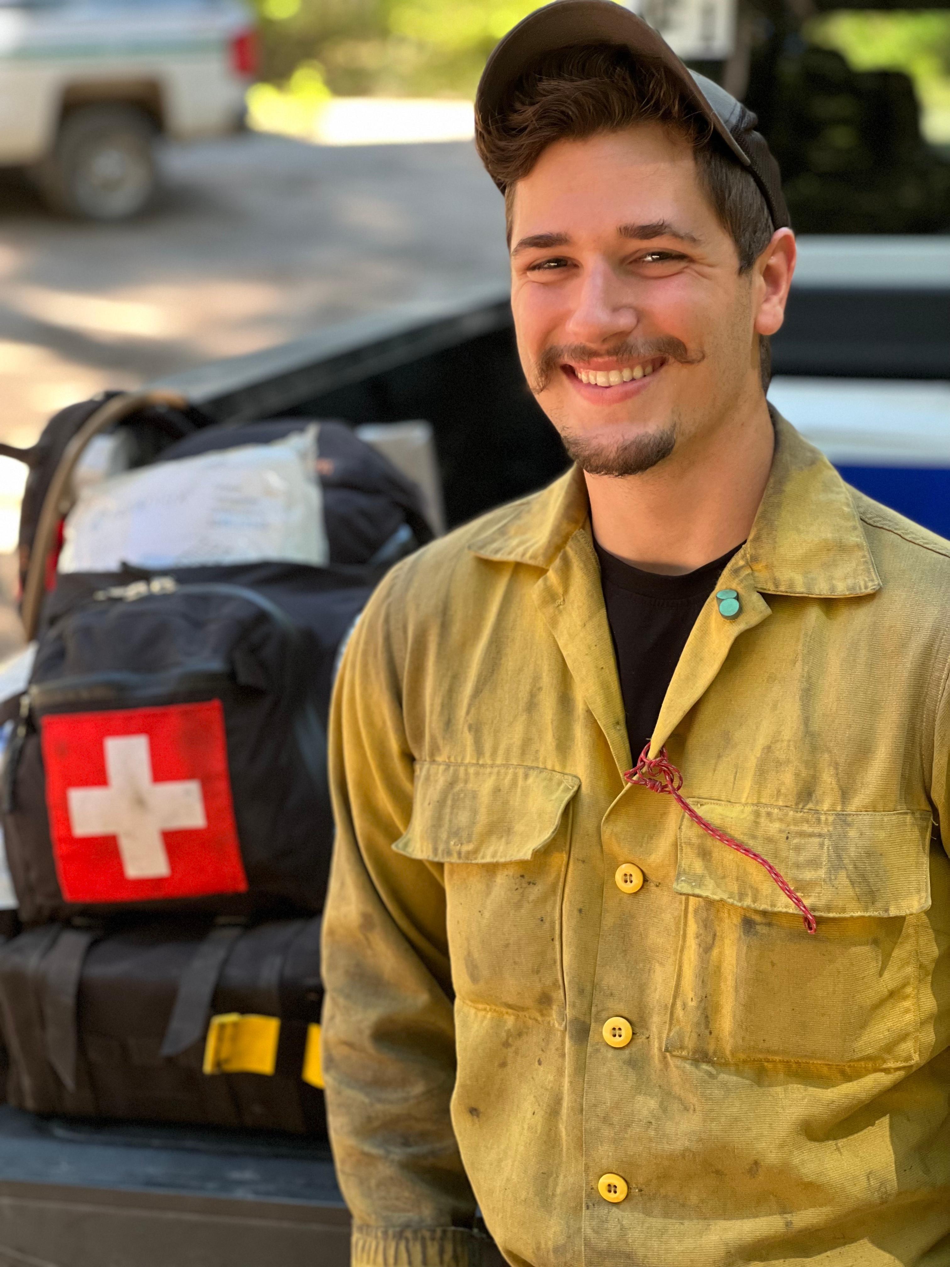 A smiling firefighter in a yellow protective shirt is seen, standing next to a pack marked with a medical symbol. His job is fireline emergency medical technician.