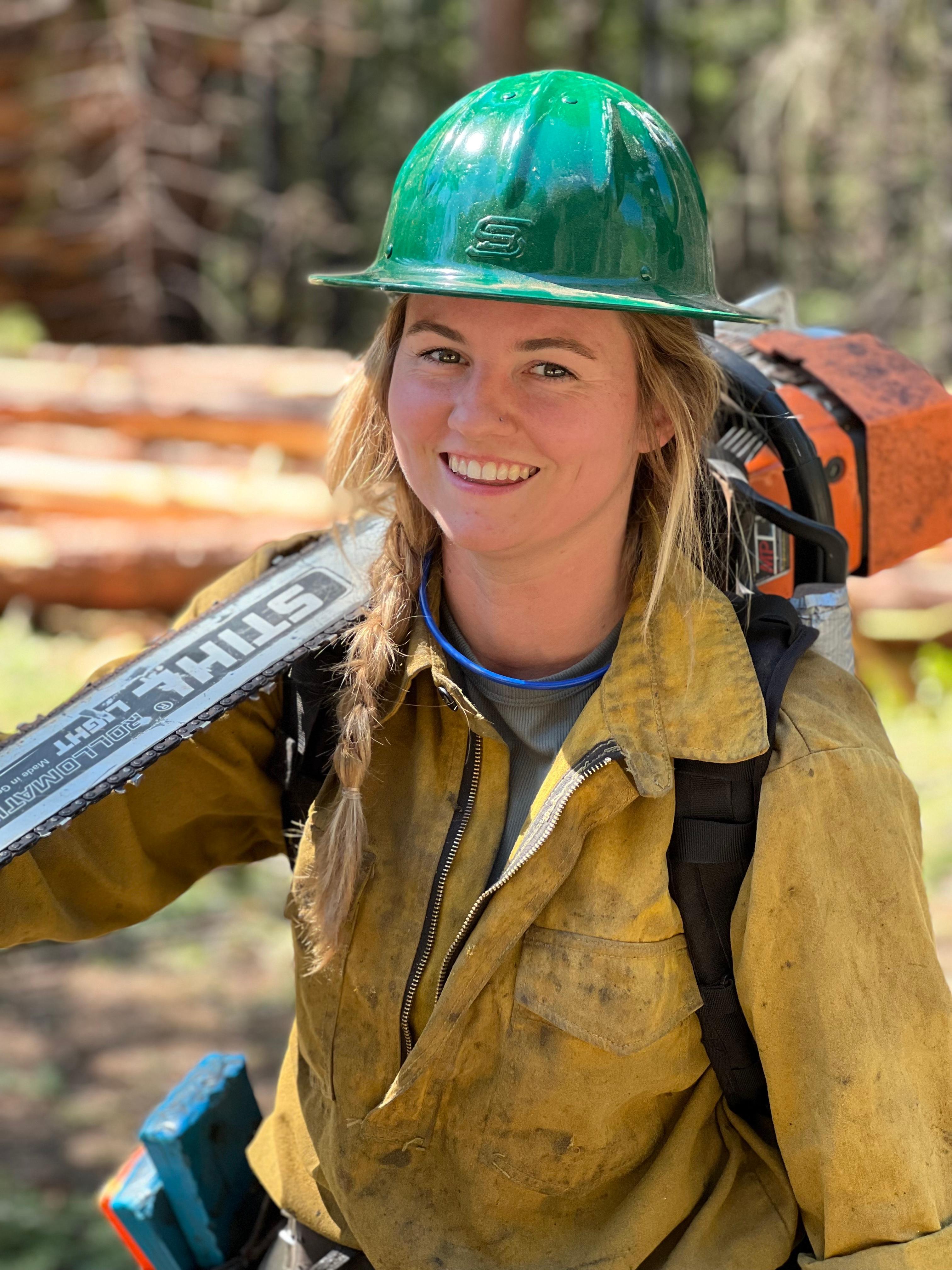 A firefighter in a green helmet is shown with a chain saw over her shoulder.