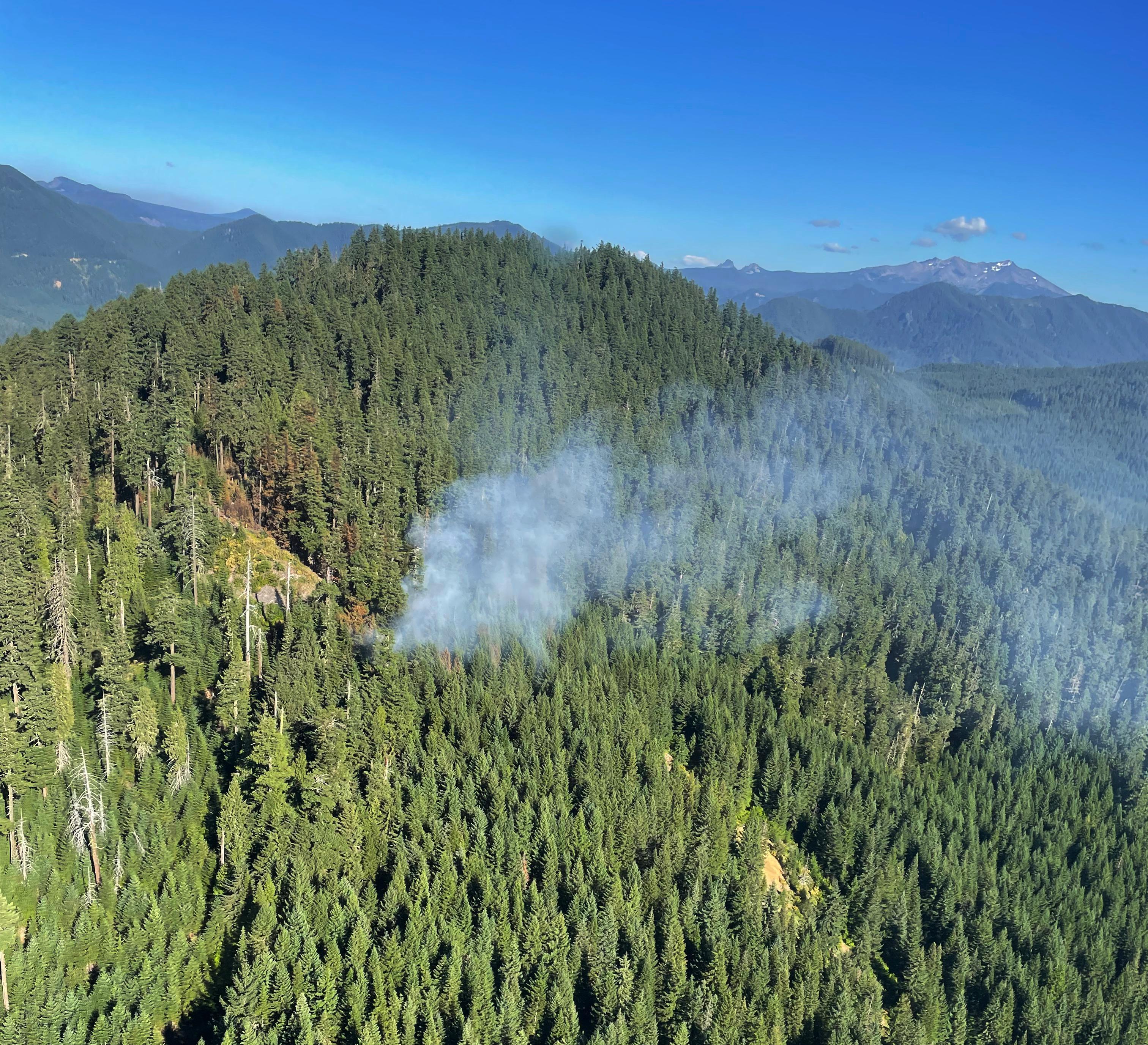 An aerial view of a small fire in a forest. Volacanic mountains with snow can be seen against a blue sky backdrop in the distance.