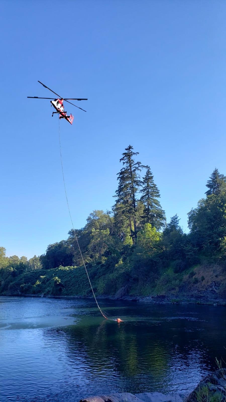 A Helicopter filling a fire suppression bucket from a large stream surrounded by trees