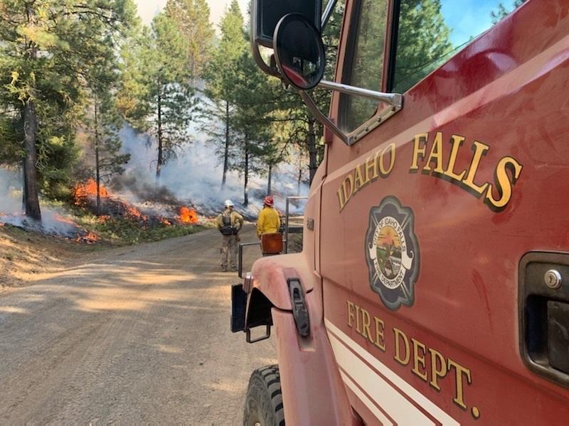 Firefighters stand along a roadside watching a creeping fire in the forest, with the Idaho Falls Fire Department Engine in the foreground.