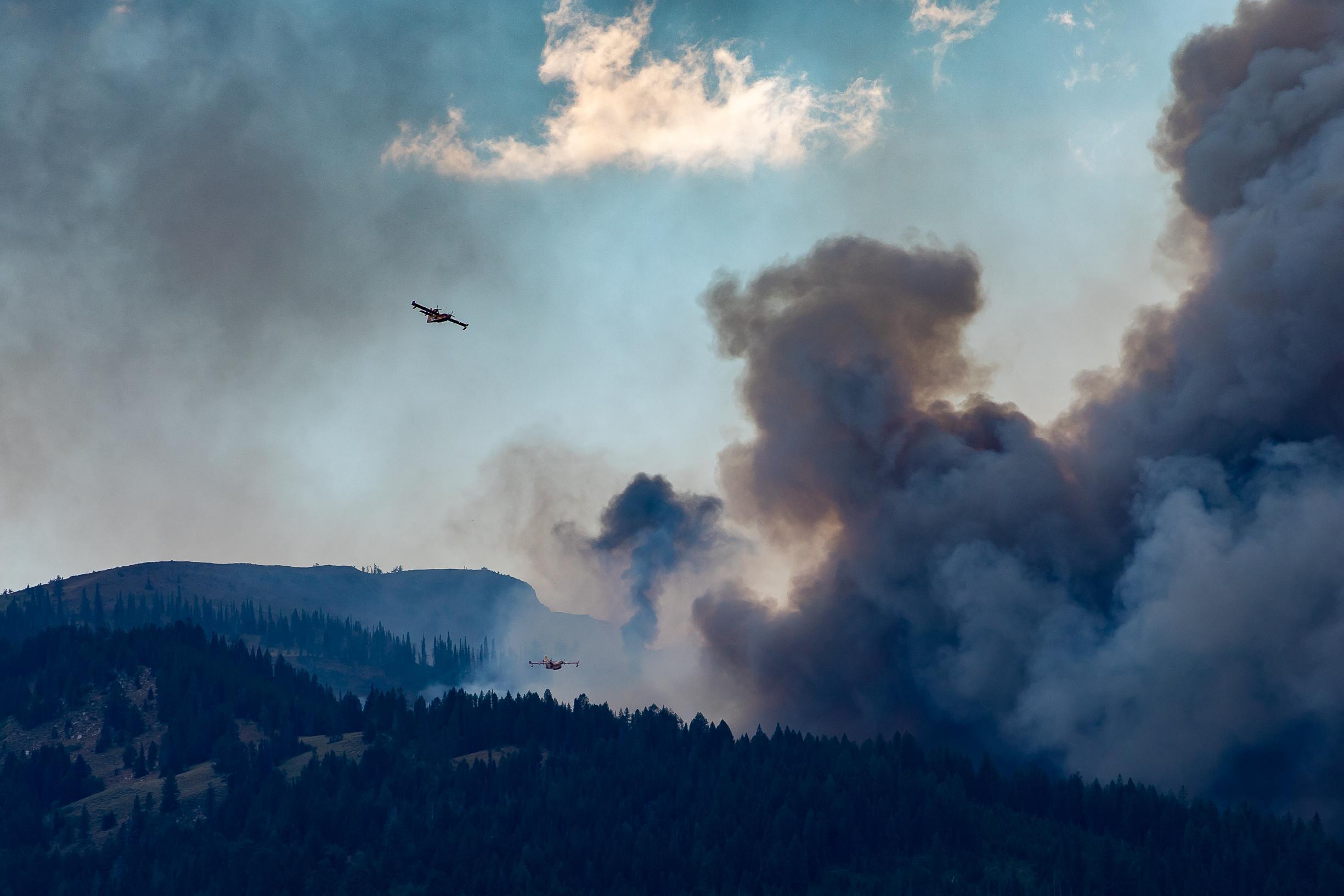 Two water carrying scooper planes fly over the mountains with smoke.