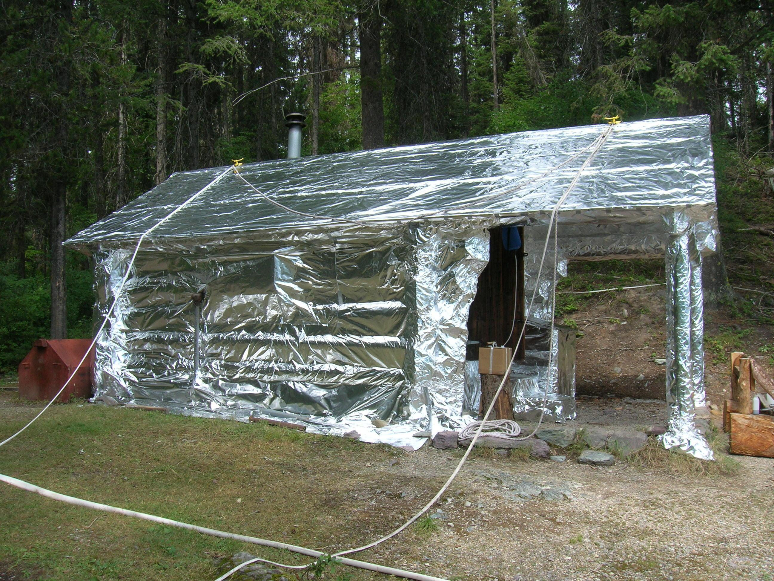 A log cabin is wrapped with silver wrap appearing similar to aluminum foil. Hoses lead to two sprinklers on the roof.