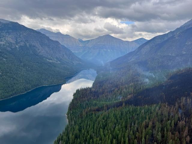 Lake surrounded by forested hillsides and rocky mountain slopes. Forest on right side of lake shows mosaic pattern of blackened trees and a few wisps of smoke.