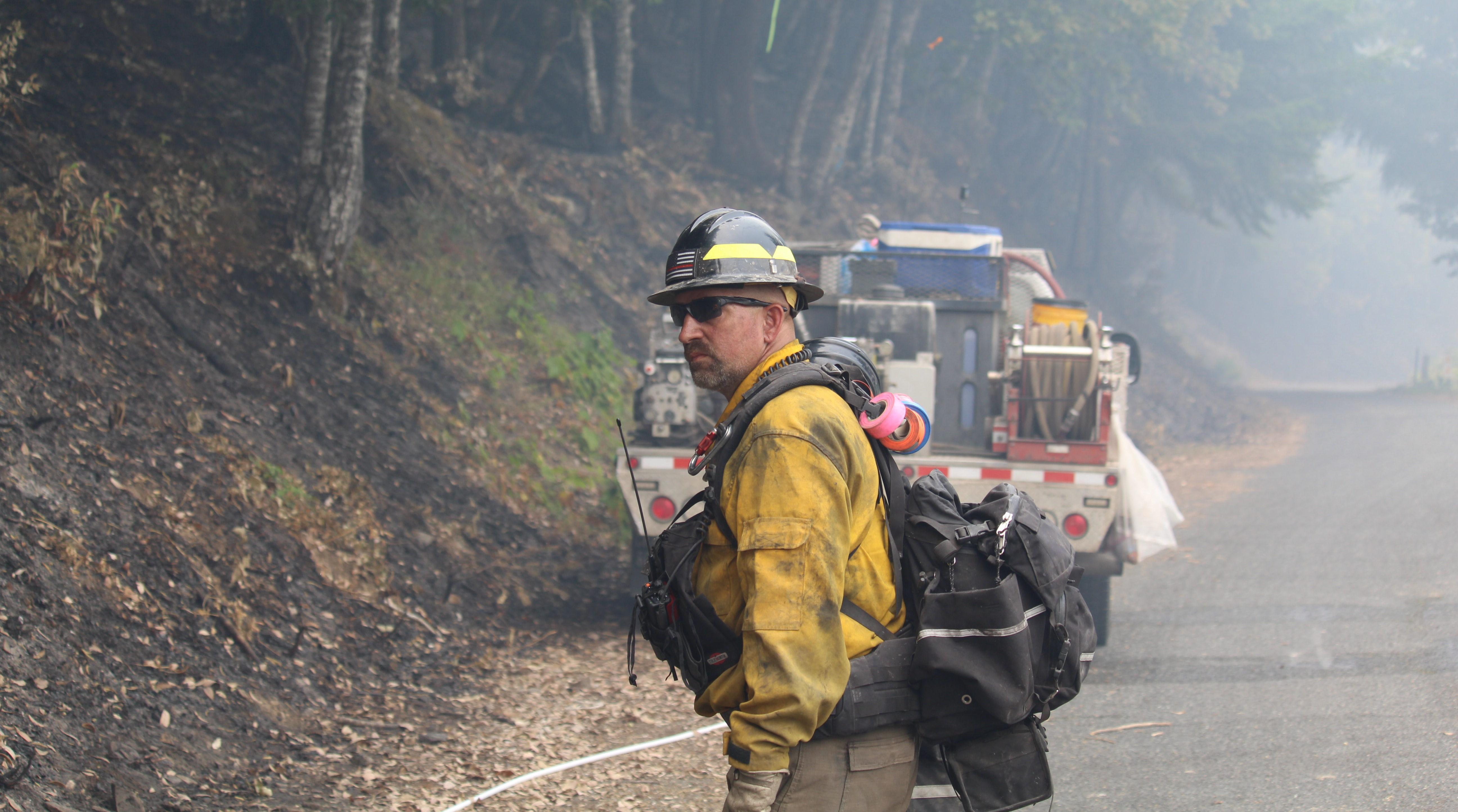 A firefighter in wildfire gear with a black helmet and sunglasses standing on a road near a steep embankment