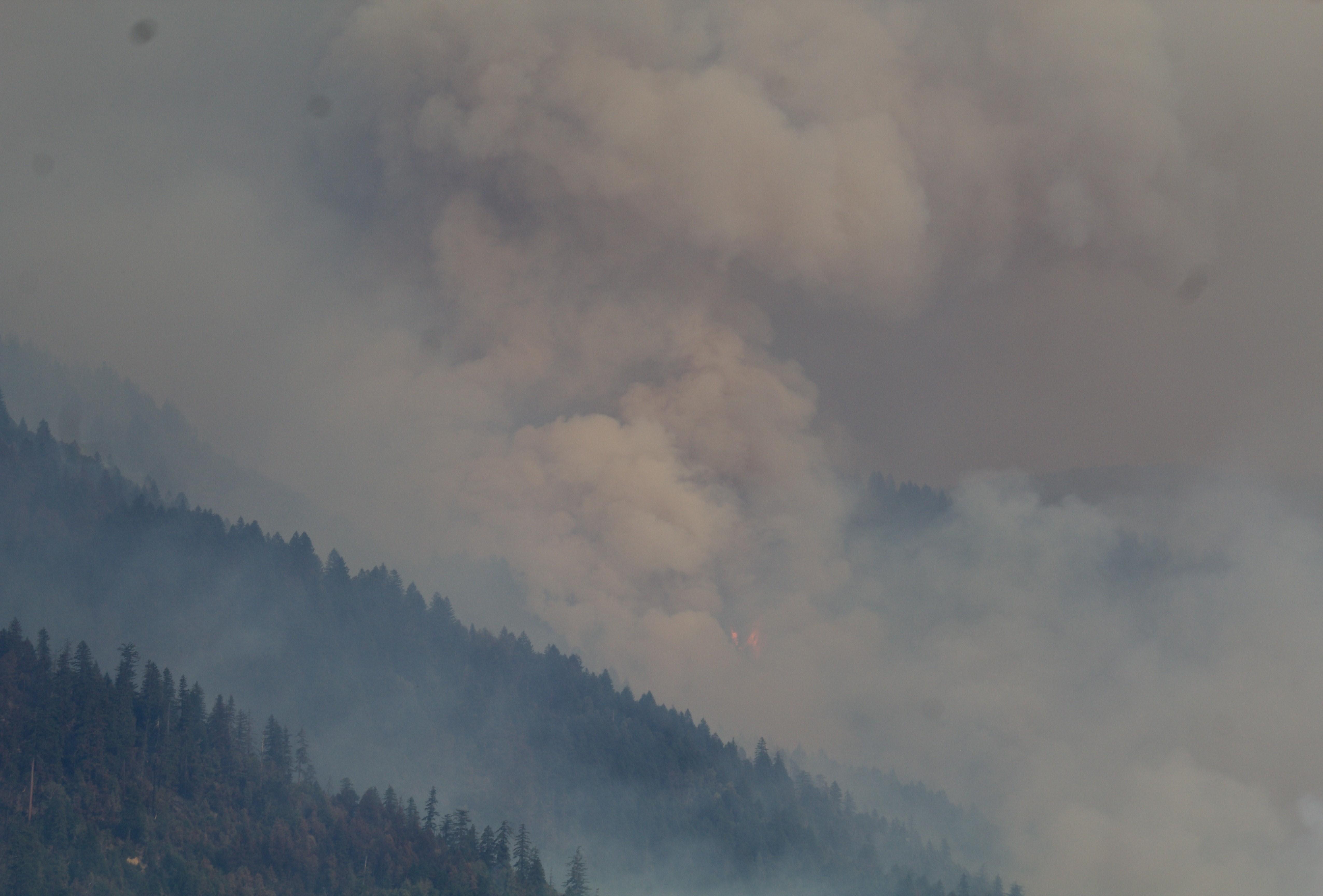 A small area of orange flames burning in a tree-covered mountain range surrounded by a thick layer of smoke