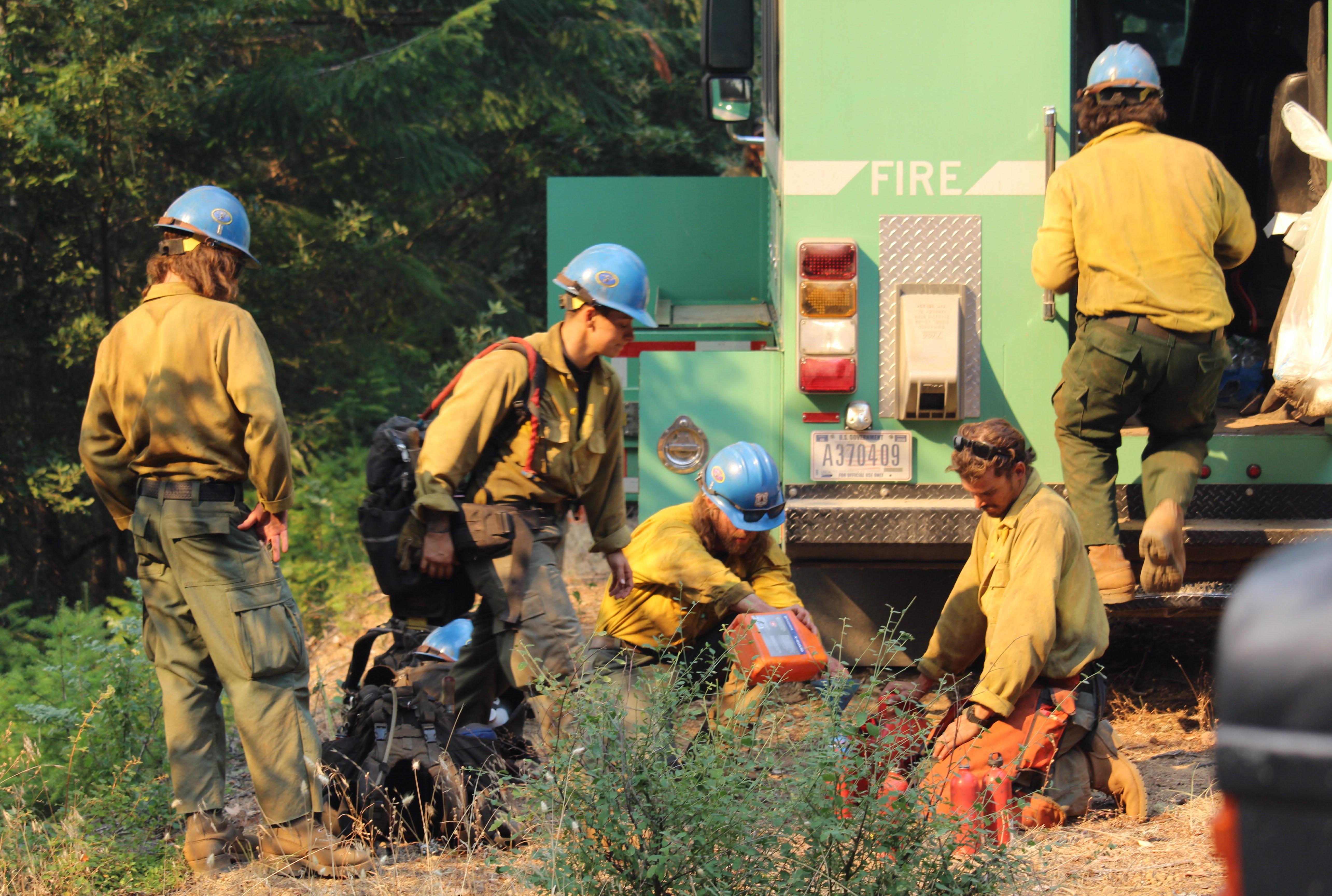 A group of hotshots in wildfire gear and blue helmets packing up various tools and equipment into large green trucks