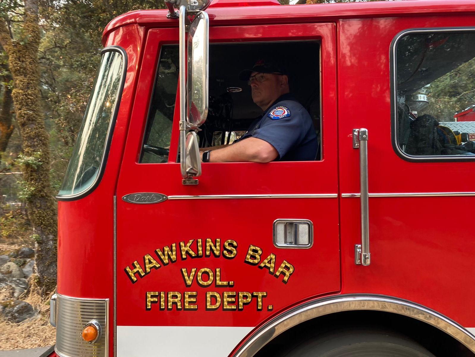 A red firetruck with gold letters reading Hawkins Bar driven by a firefighter in a blue uniform