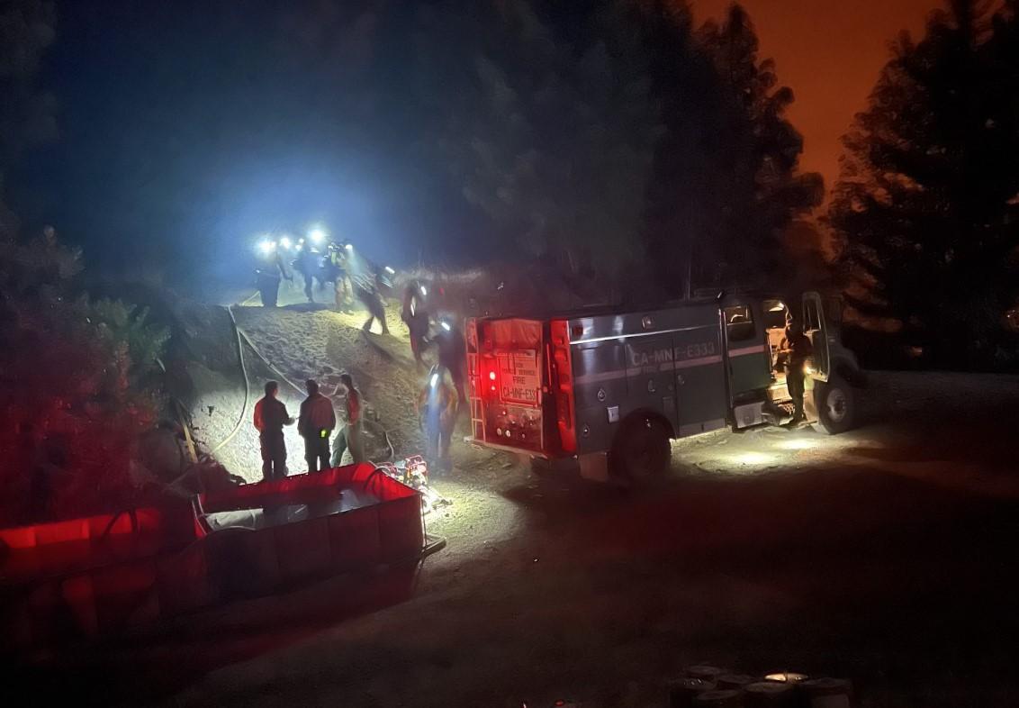 A group of firefighters in wildfire gear, with lit headlamps hike down a hill near a green fire engine drawing water from a square tank