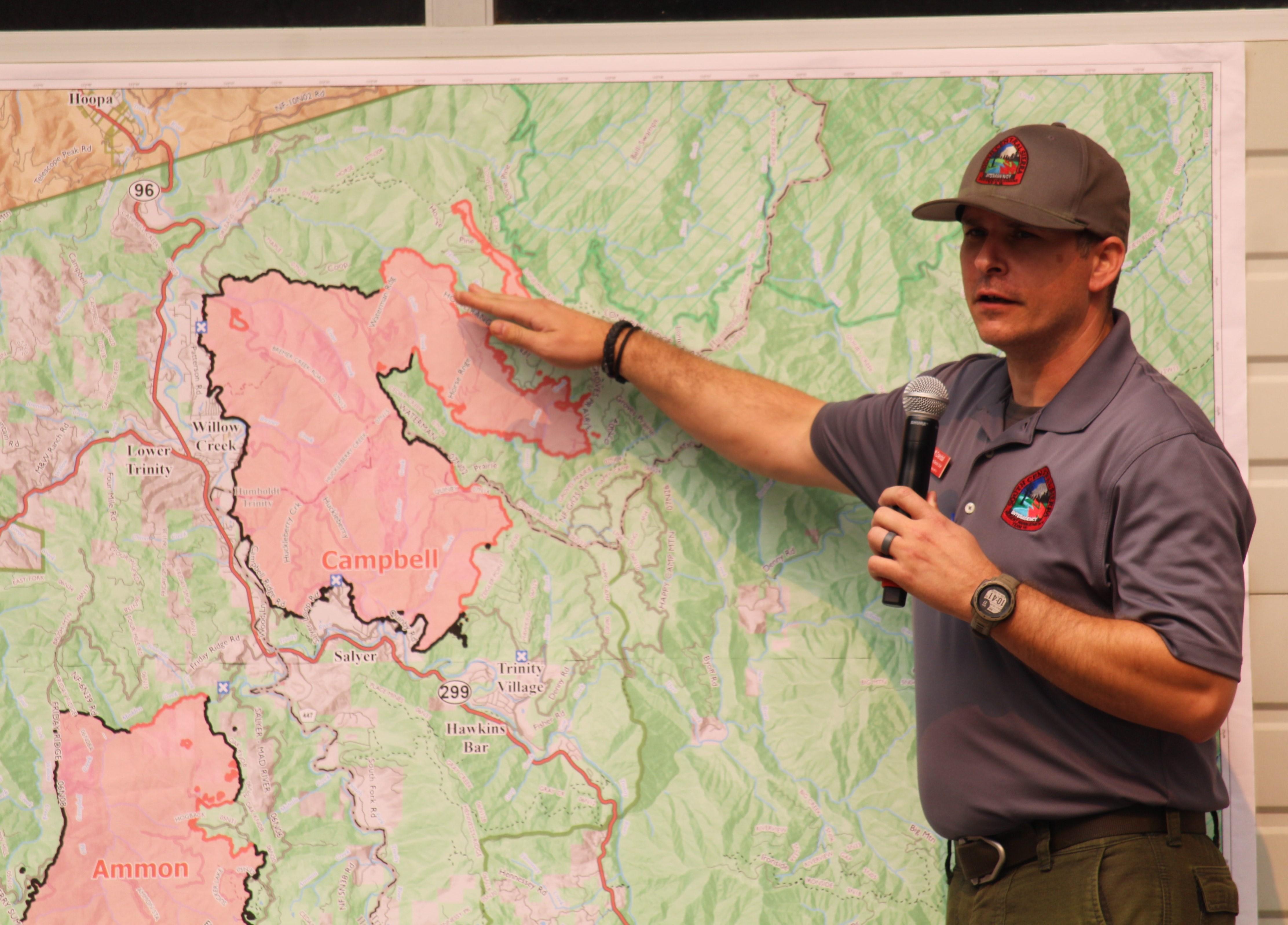 A man in a grey shirt and hat gestures to a map as he speaks into a microphone