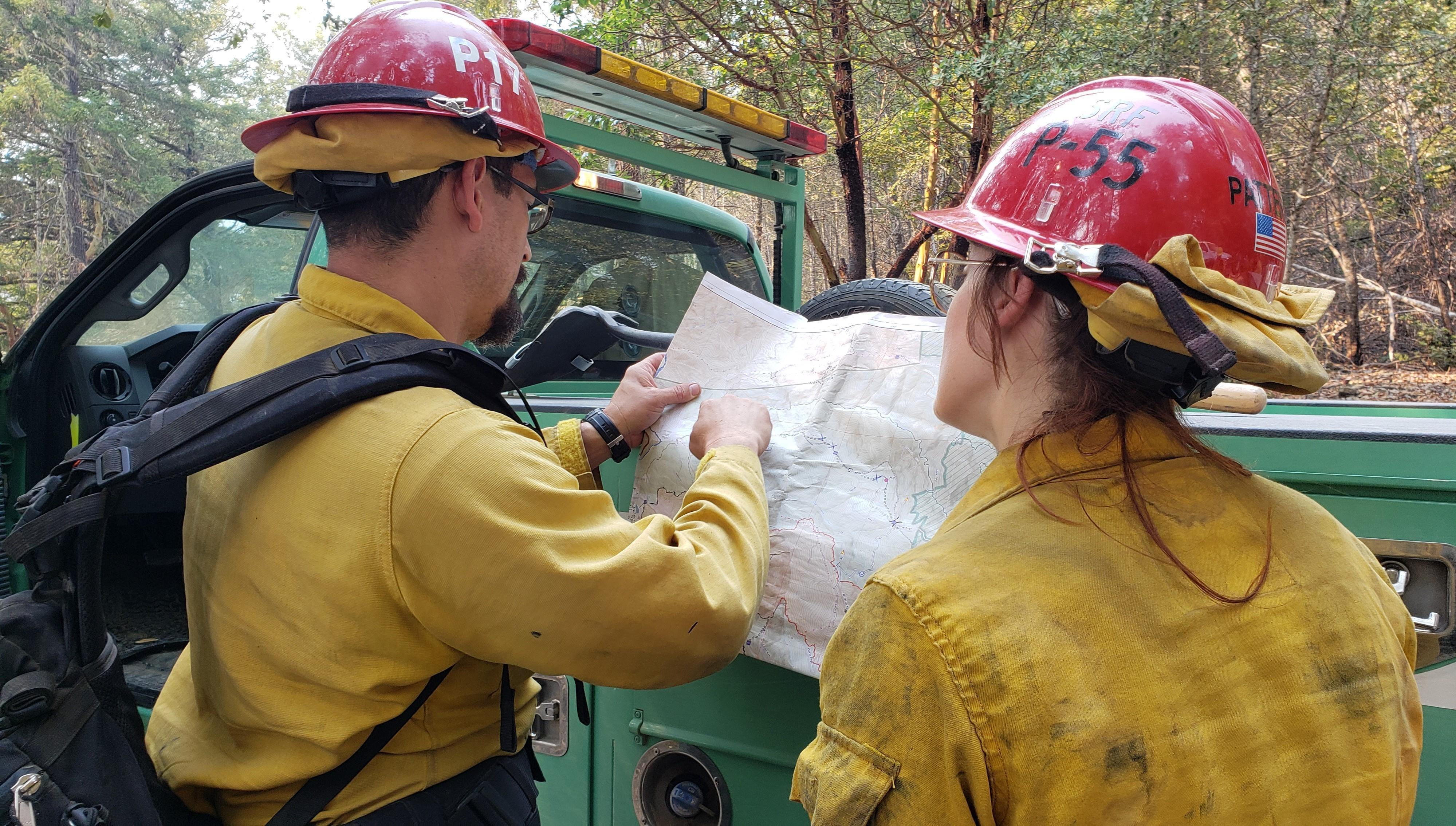 Two firefighters in wildland gear and red helmets study a map against a green truck