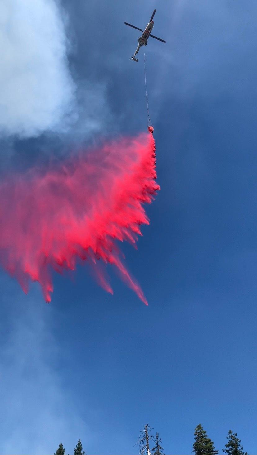 A white and green helicopter drops red fire retardant from an orange bucket