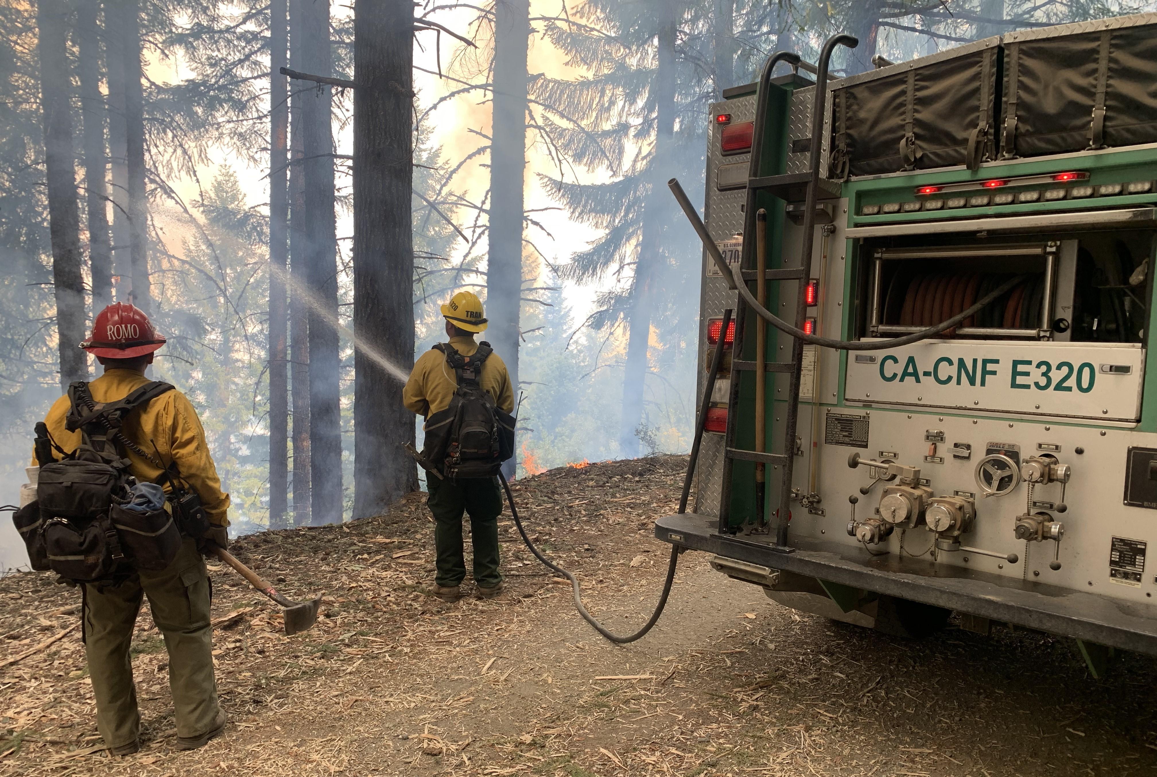Two Firefighters in green pants and yellow shirts, red and yellow hats standing next to engine, using hose along the control lines on Lone Pine Ridge and to the north of the fire. 