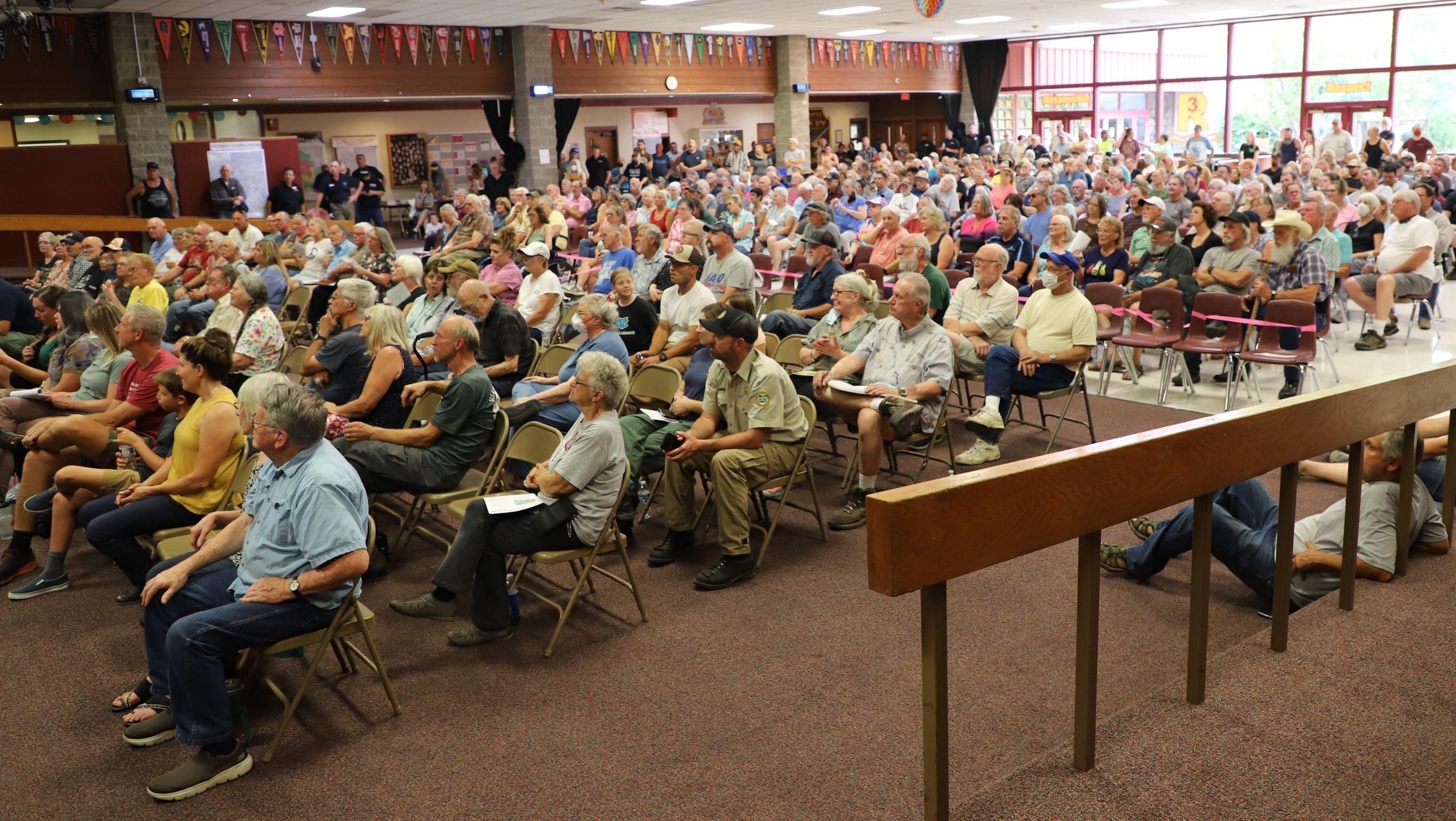 A large audience of people are sitting in chairs and on the floor, listening to the speakers at the community meeting held August 30 in Grants Pass.