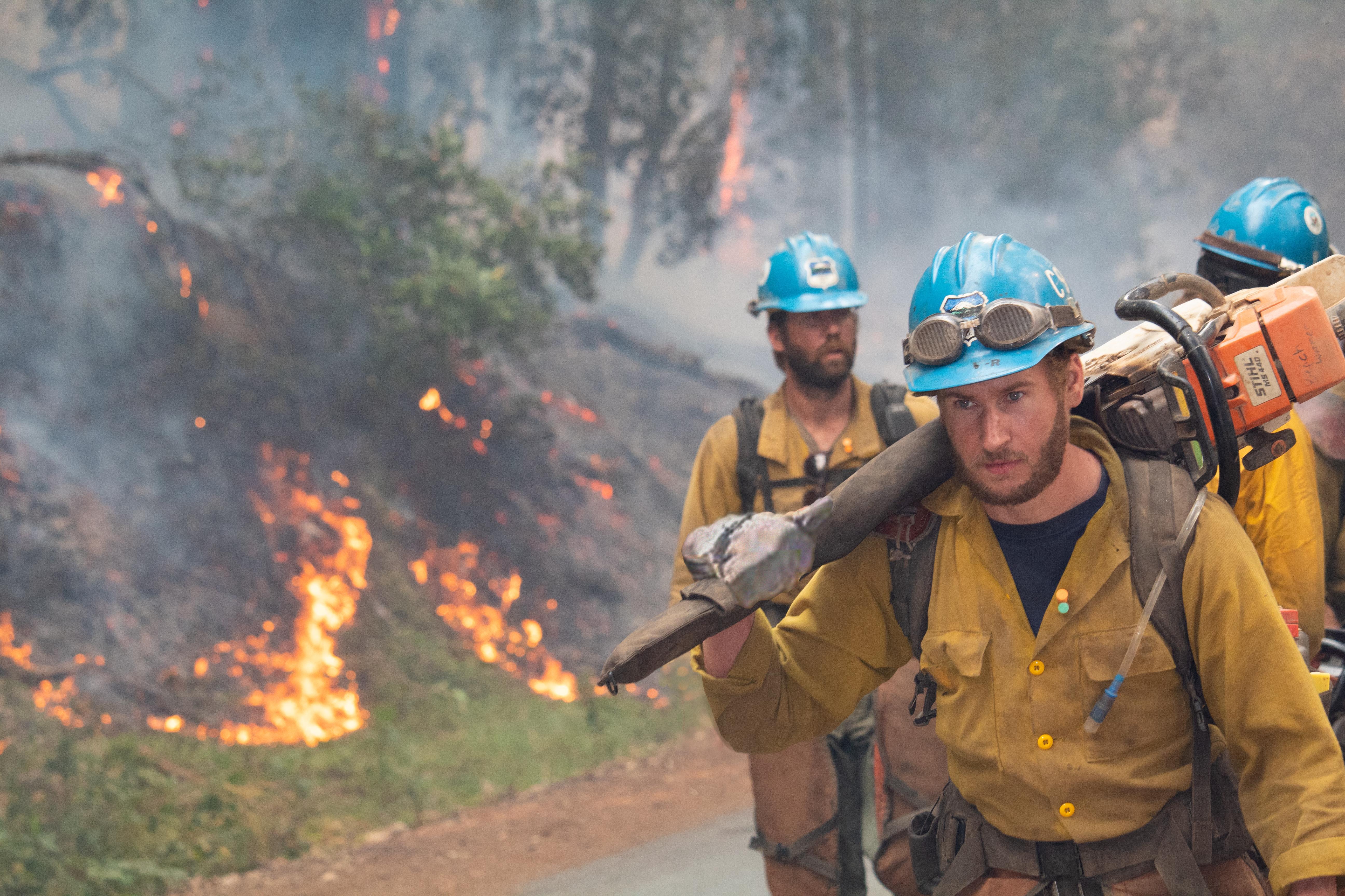 Three Crew members walking along road facing camera, wearing blue hardhat, fire on left on hill.