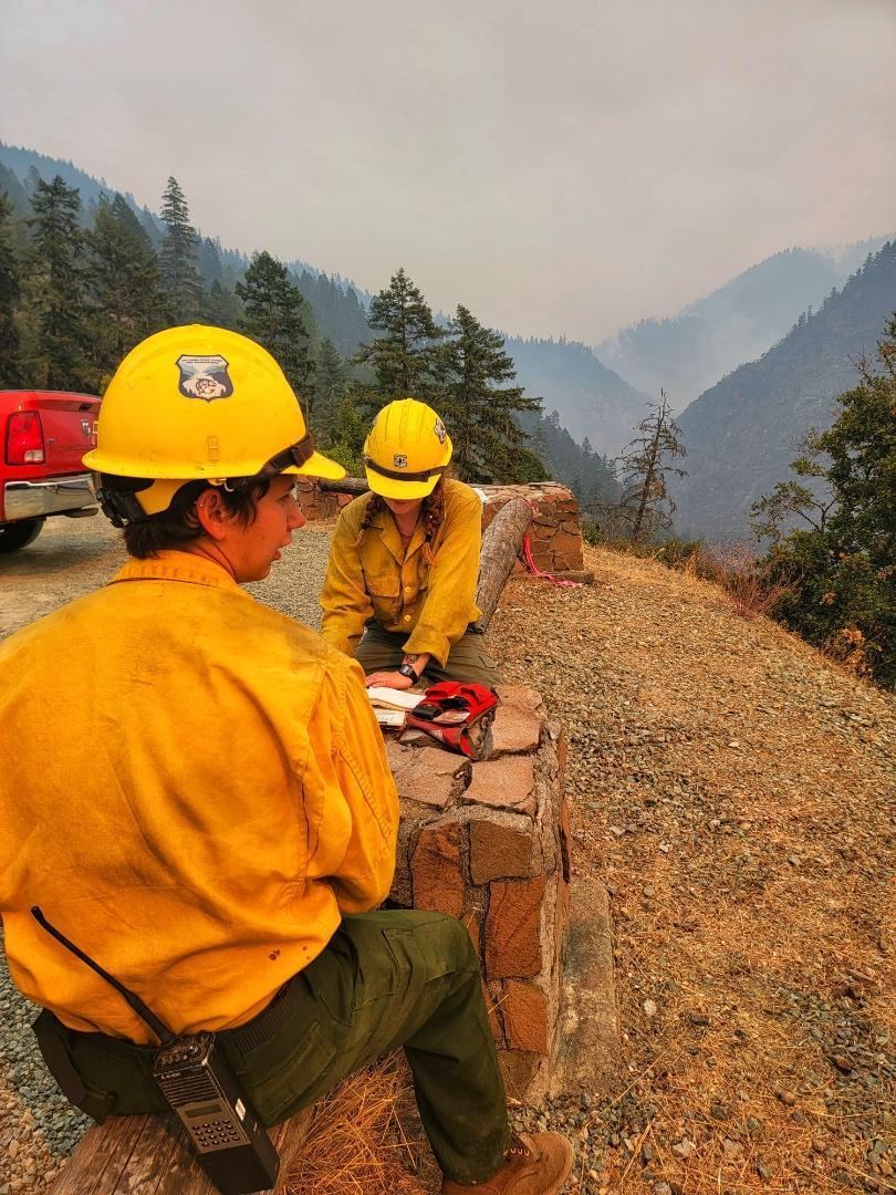 Two firefighters sit at the viewpoint at Rainie Falls, looking south across the Rogue River at the Rum Creek Fire area. They are watching for flare-ups and changes in fire activity.